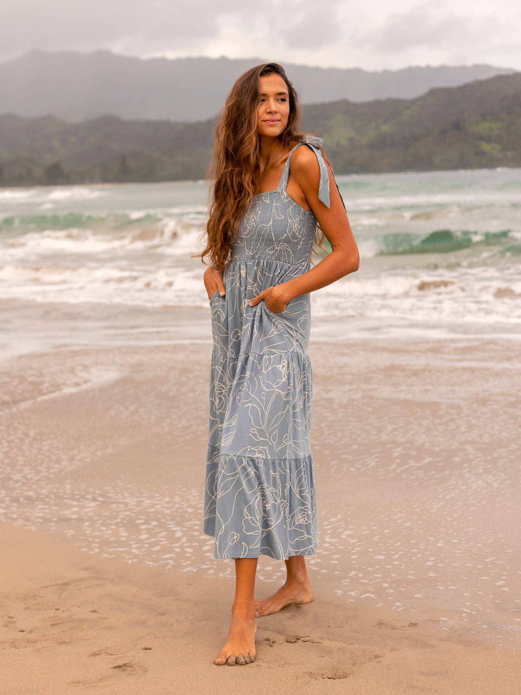 Woman in Evie Smocked Dress in Garden Rose, a light blue maxi with white floral print, smocked bodice, and ribbon ties, standing on a beach.