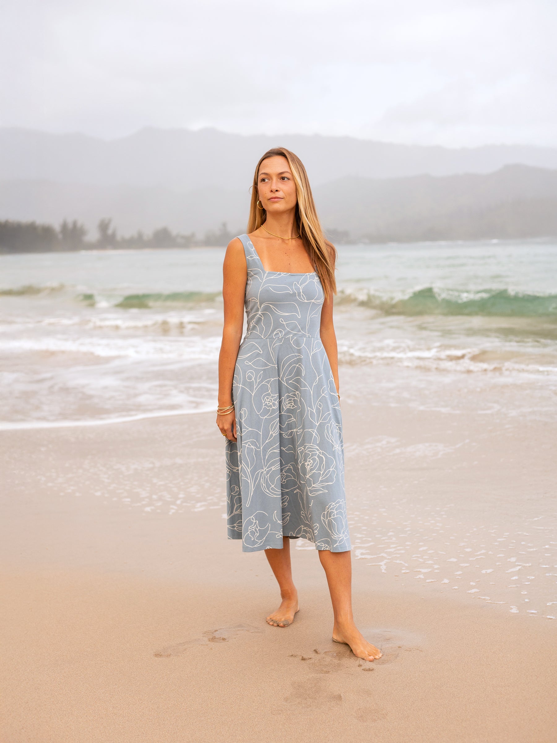 Woman in a blue organic cotton Lily Dress with white garden rose print, square neckline, and midi skirt, standing on a beach.