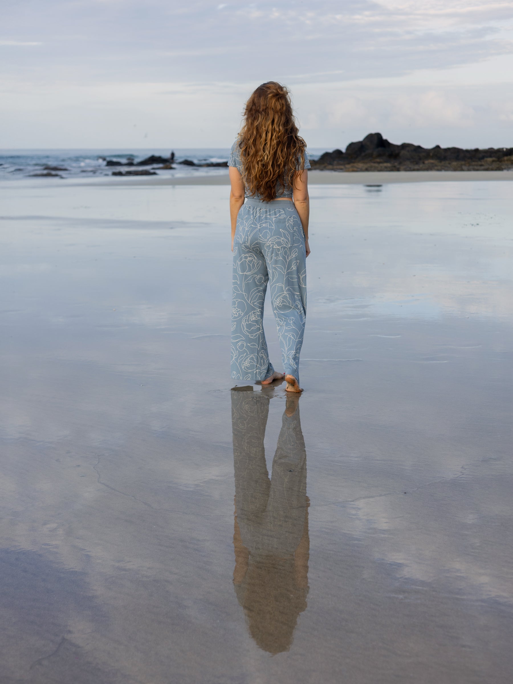 Woman from behind wearing light blue wide-leg organic cotton pants with white rose print on a wet beach.