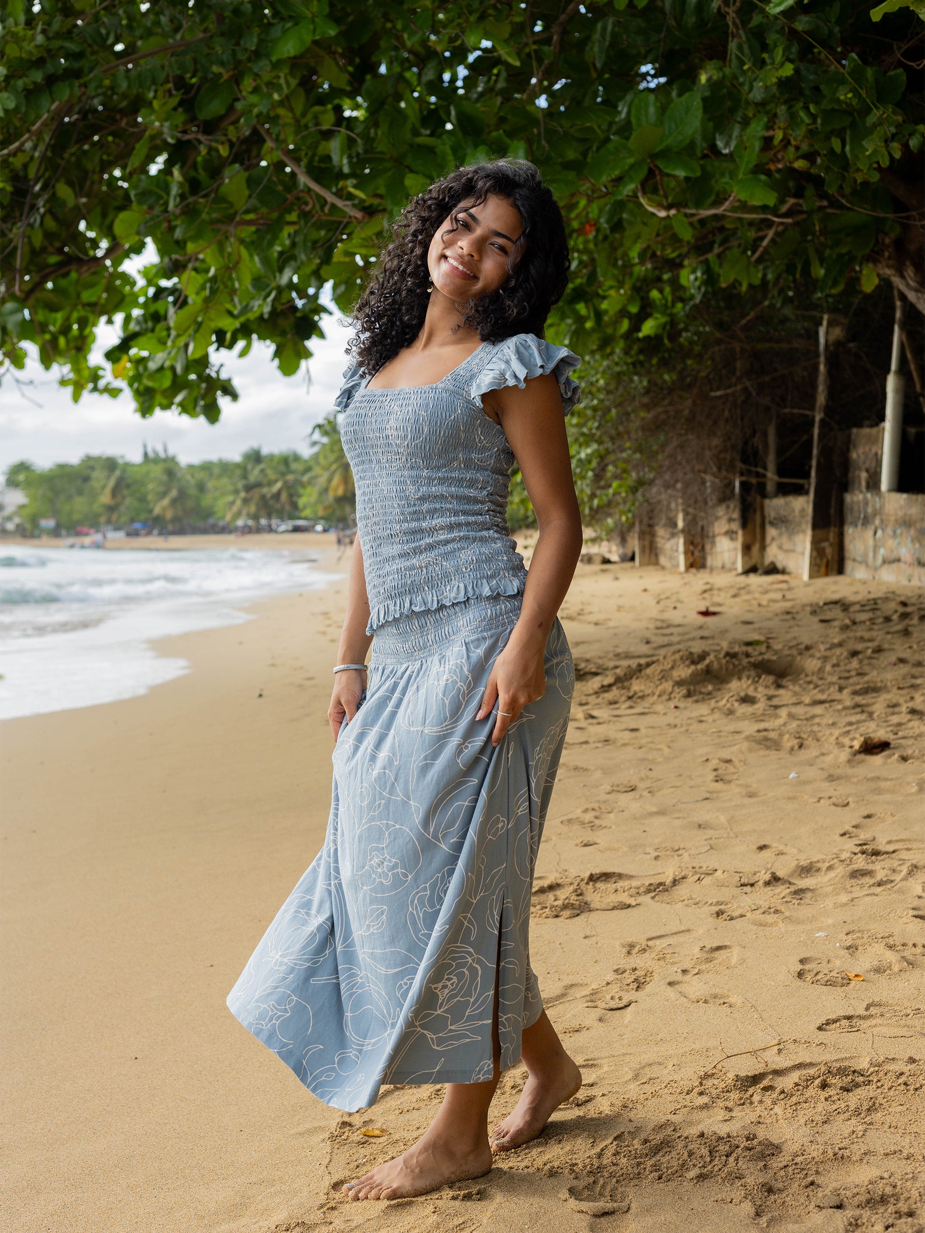 Woman in a blue smocked top with flutter sleeves and matching skirt with white rose print on a sandy beach.