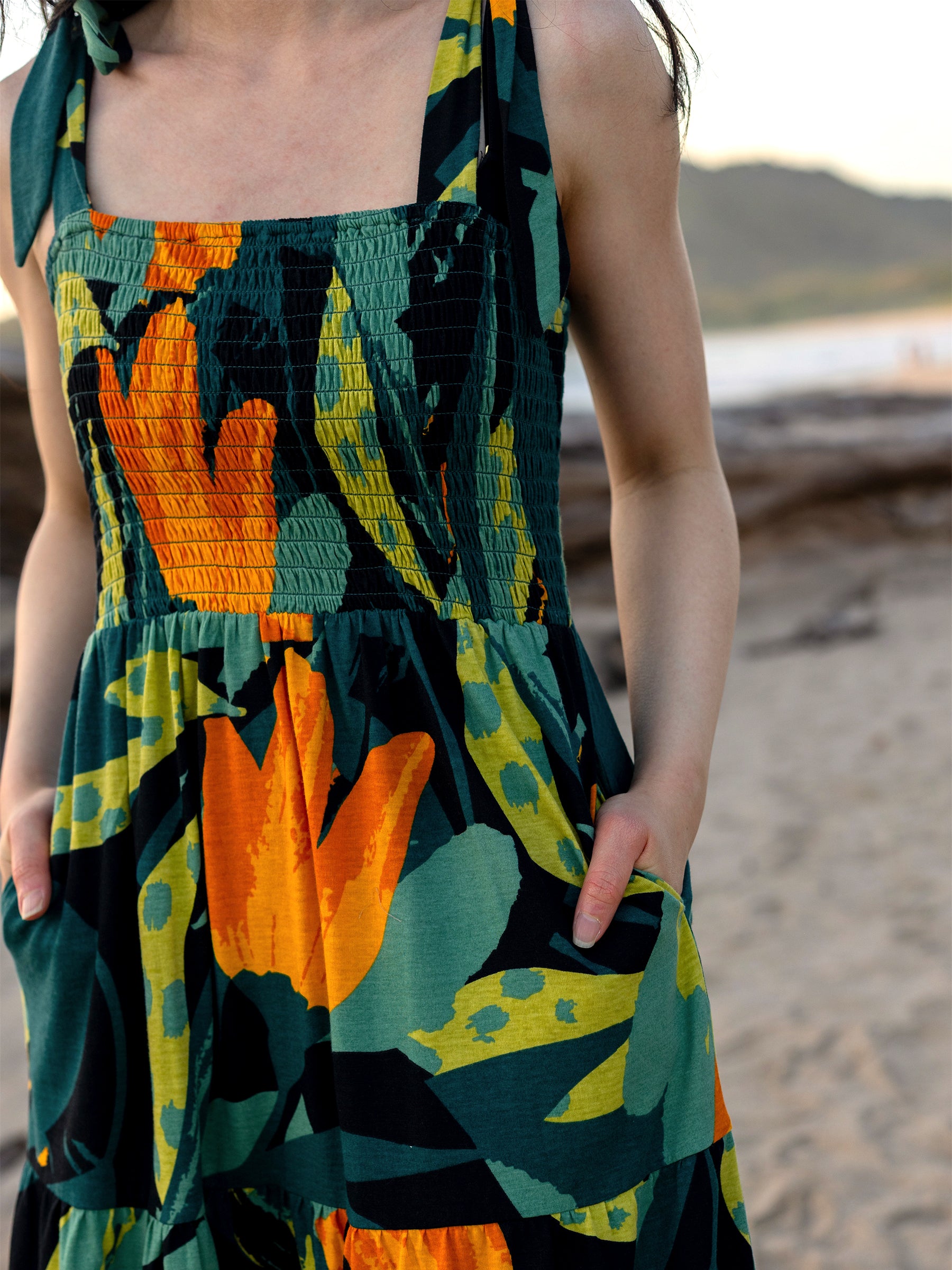 Evie Smocked Dress in organic cotton, featuring a vibrant green, orange, and yellow tropical print, smocked bodice, and pockets.