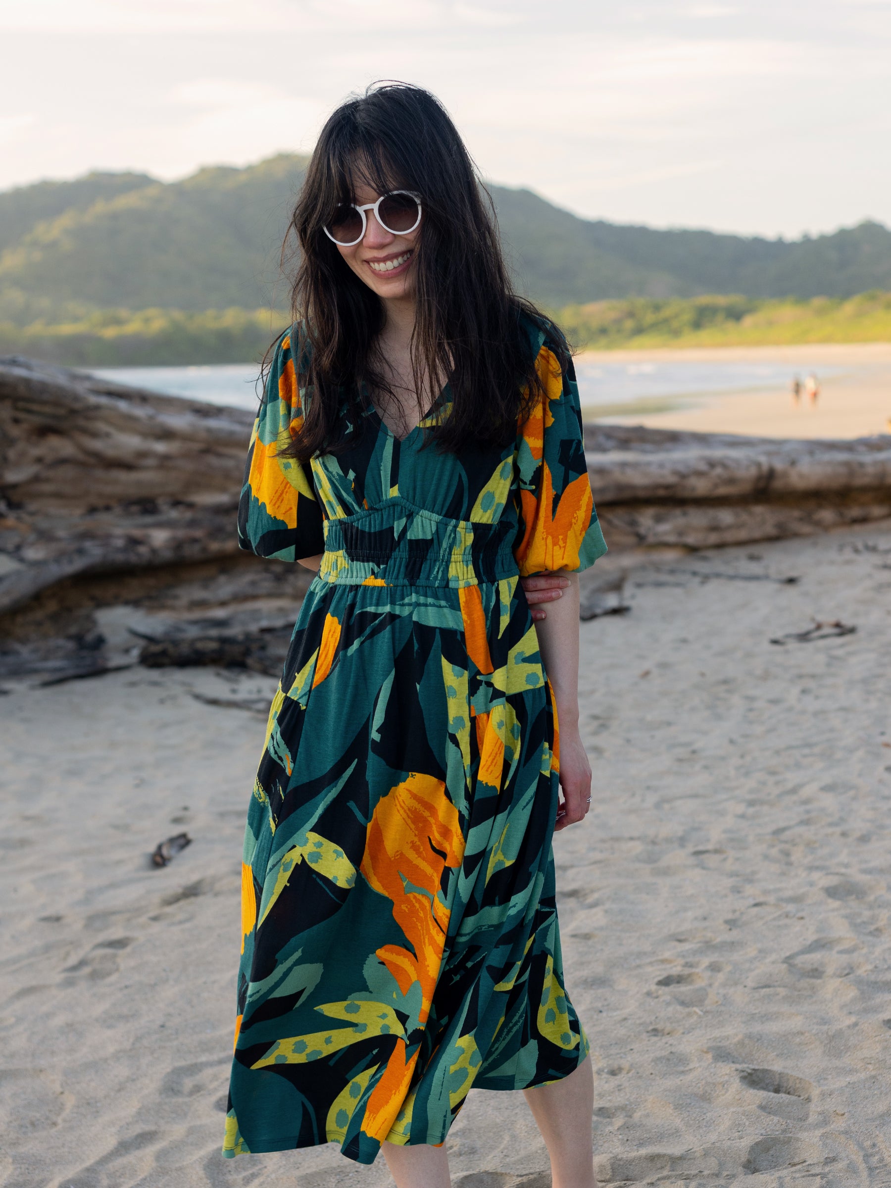 Woman in Ophelia Dress: green, orange, and black floral midi dress with V-neck, puffed sleeves, and smocked waist on a beach.
