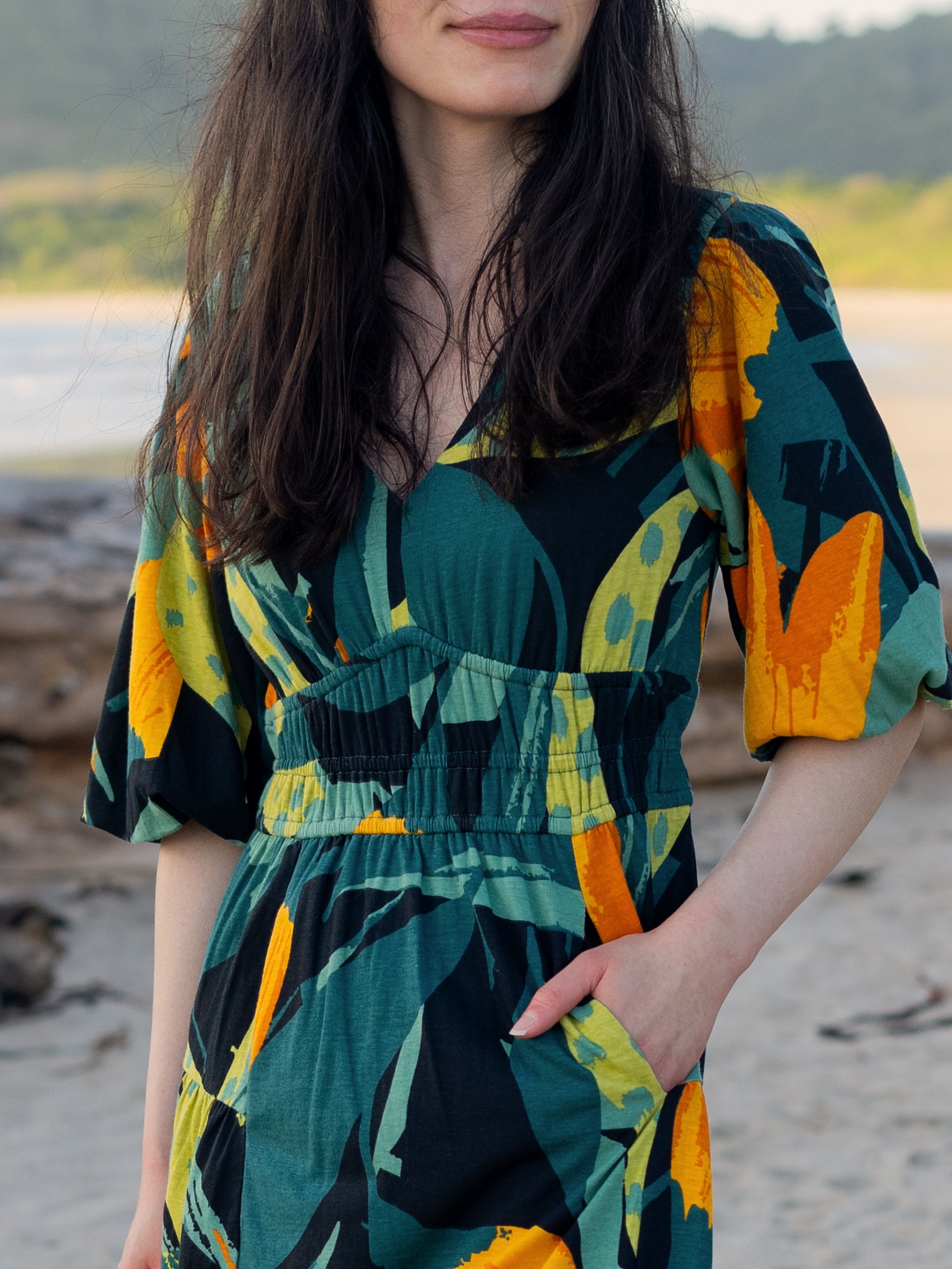 Ophelia Dress: Woman in a green, yellow, and black tropical print midi dress with puffed sleeves and V-neck.