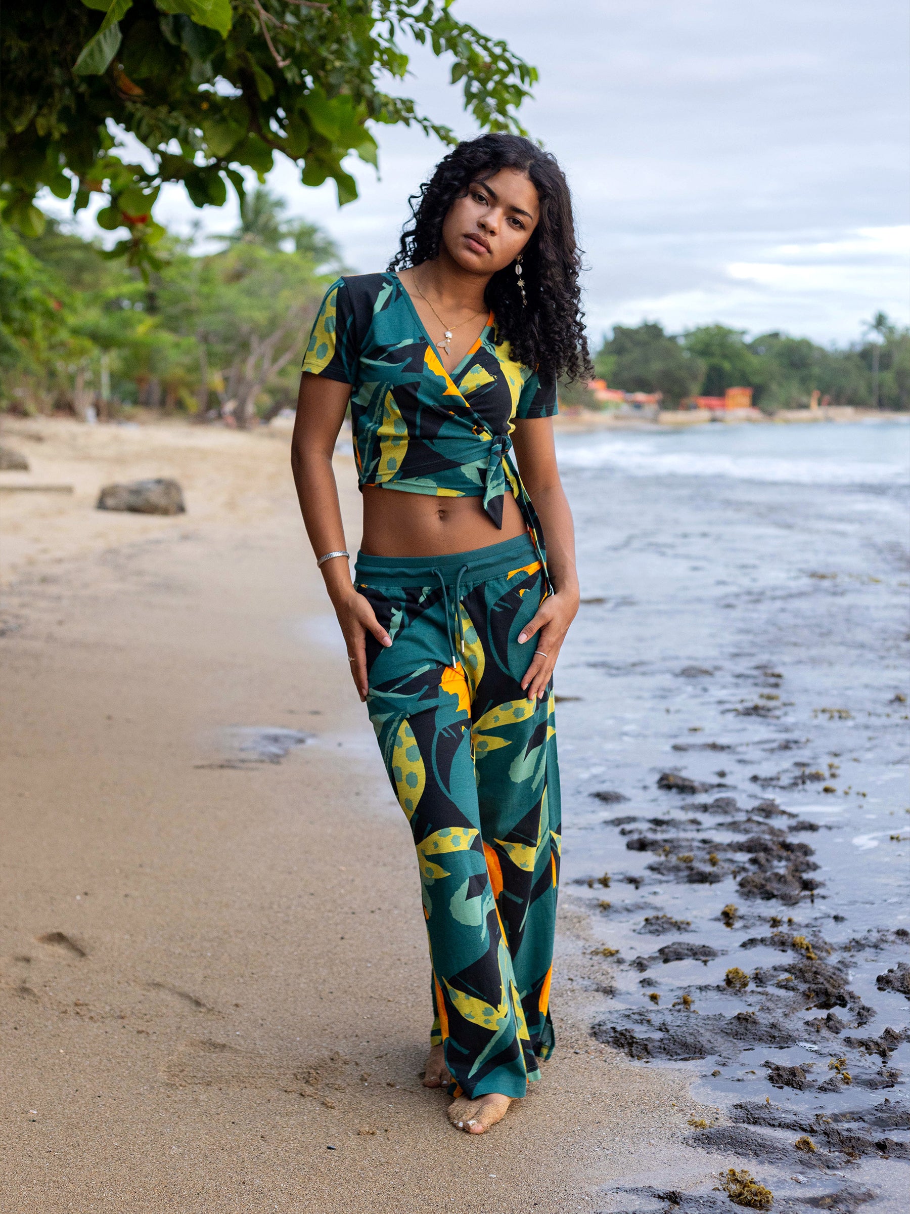 Woman in a green and yellow Hanalei wide-leg pant set with a wrap top, standing on a sandy beach.