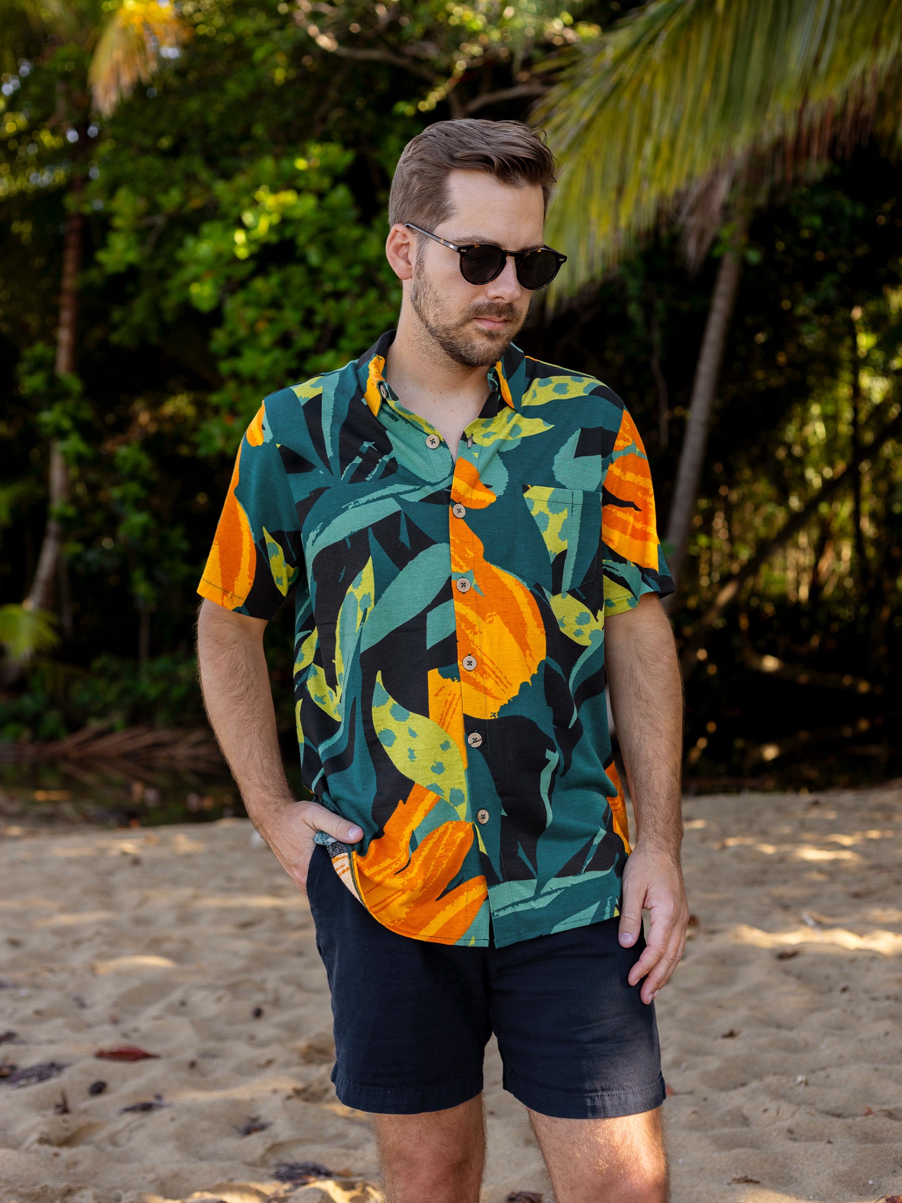 Man in Rincon Shirt, a green, orange, and black tropical print organic cotton button-up, on a sandy beach.