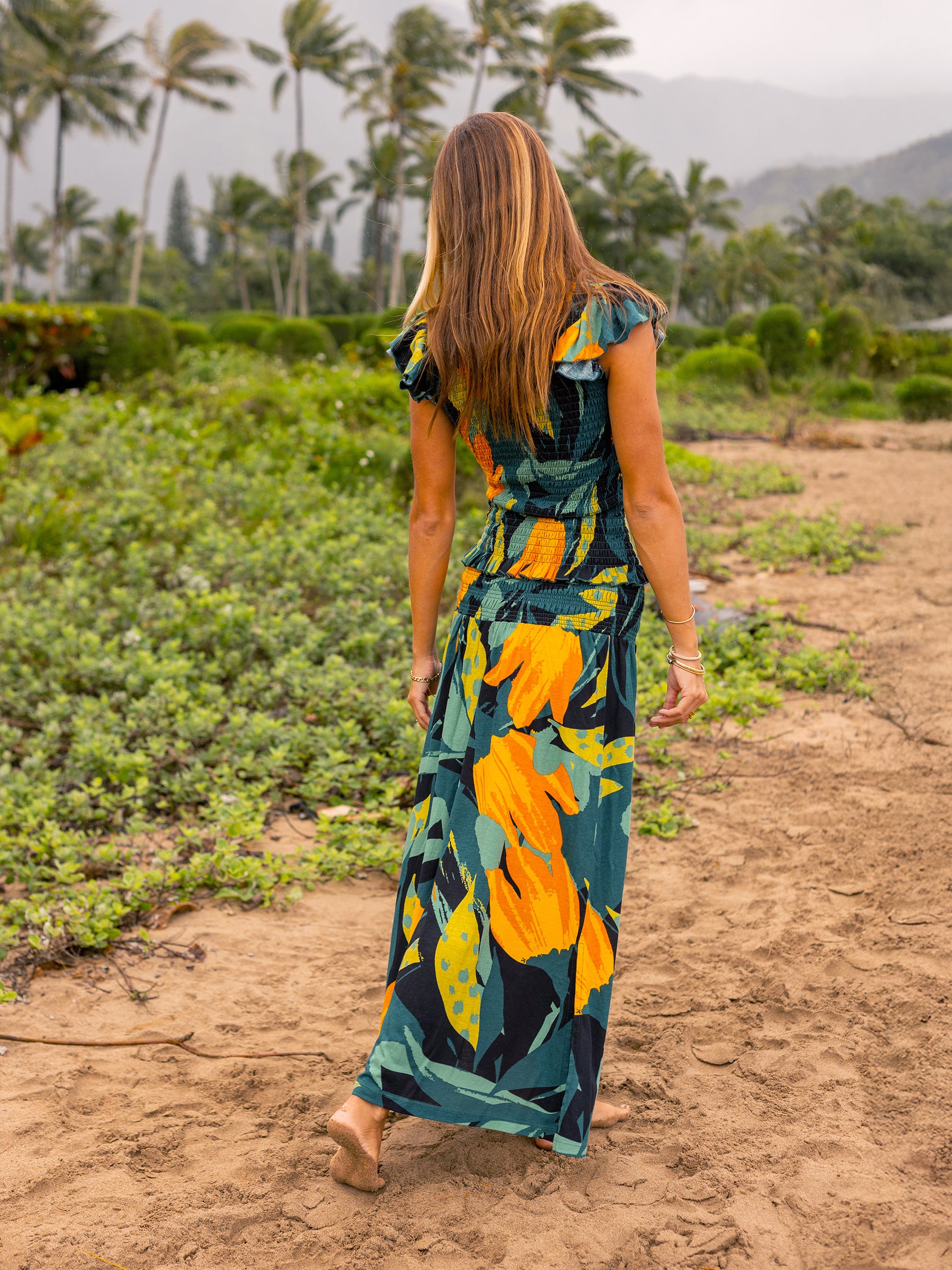Woman in a Hanalei smocked top and matching maxi skirt with a vibrant floral print, walking on a beach.