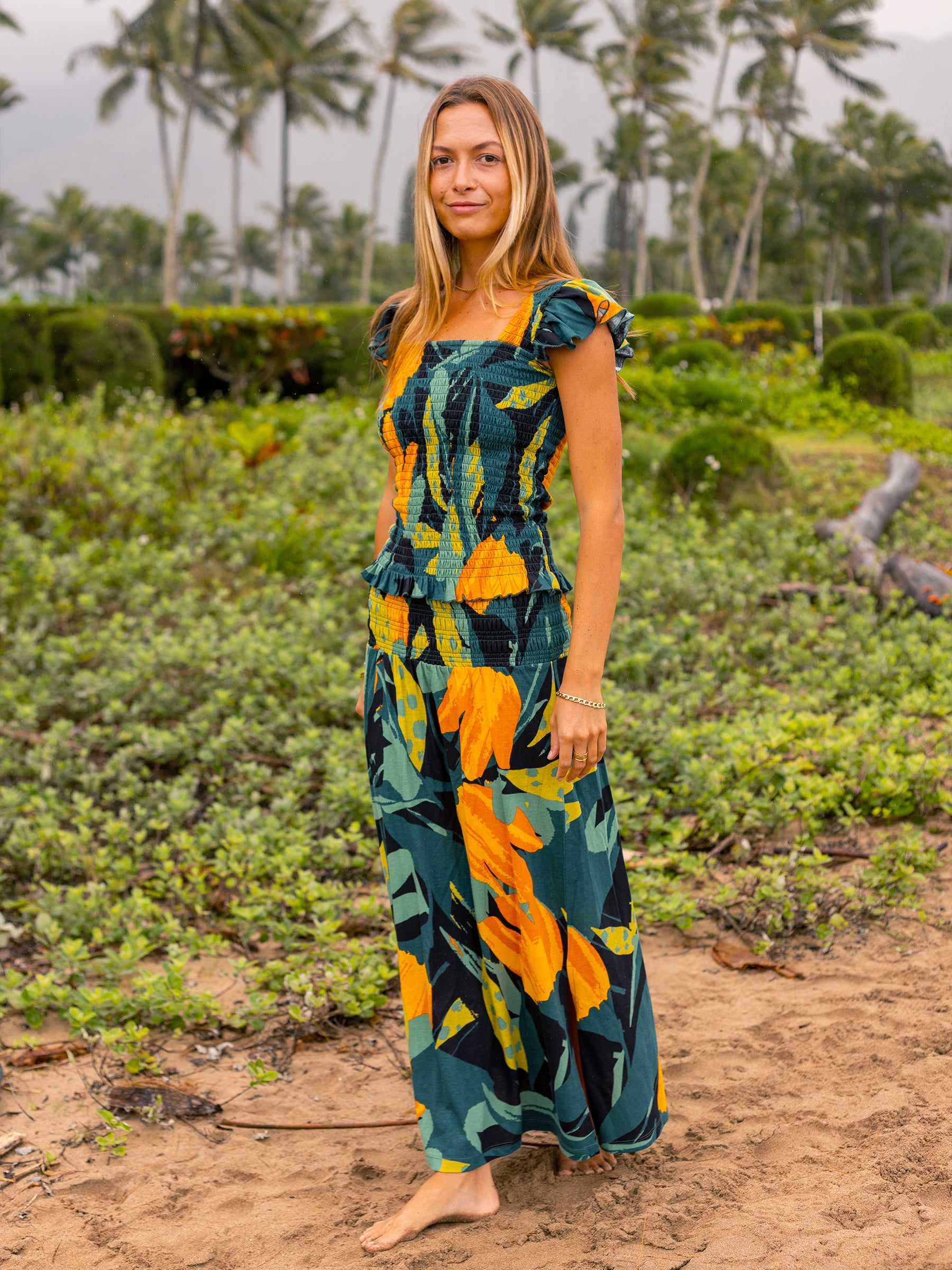 Woman in a Hanalei smocked top and matching pants with green and golden floral print, flutter sleeves, and ruffled hem.
