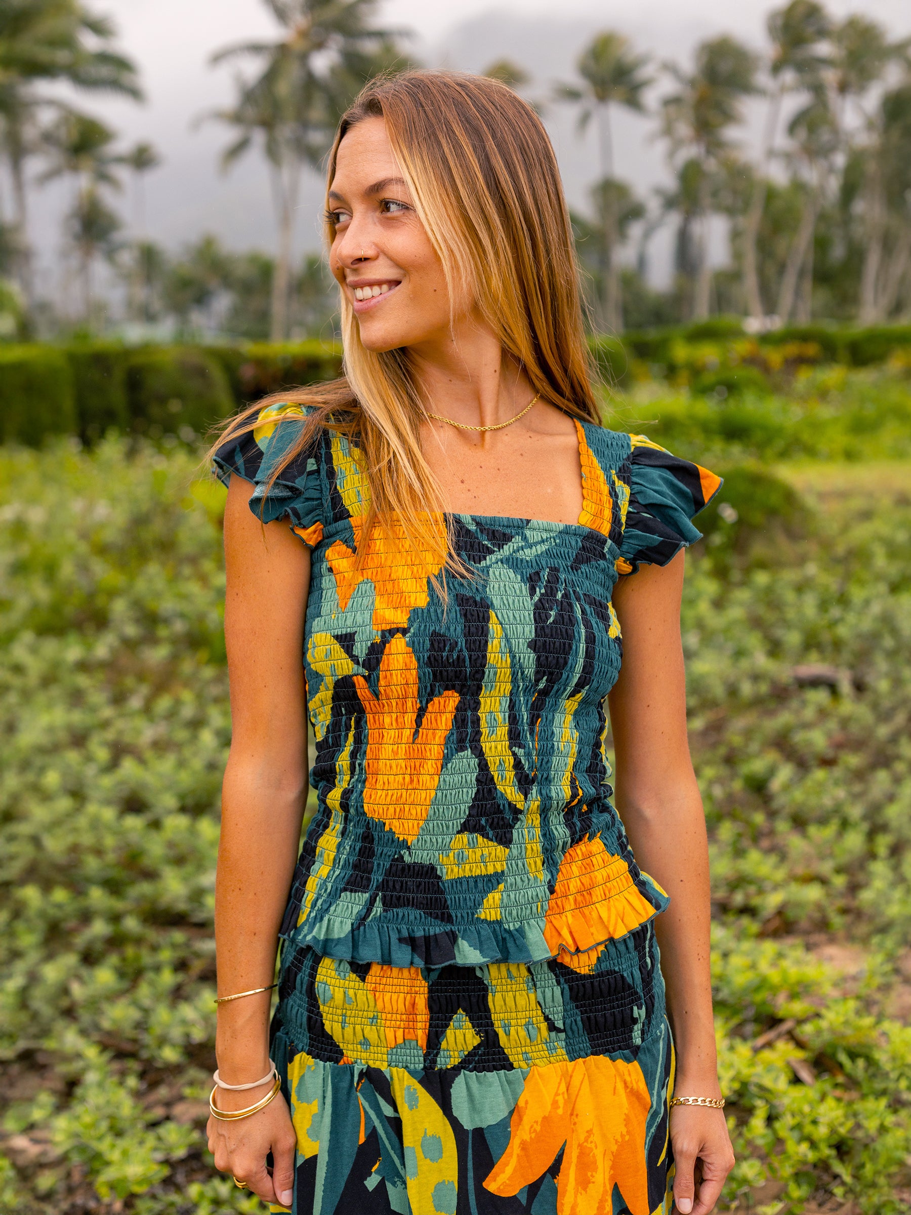 Smiling woman in a Hanalei smocked top with a vibrant green and golden floral print, square neckline, and flutter sleeves.
