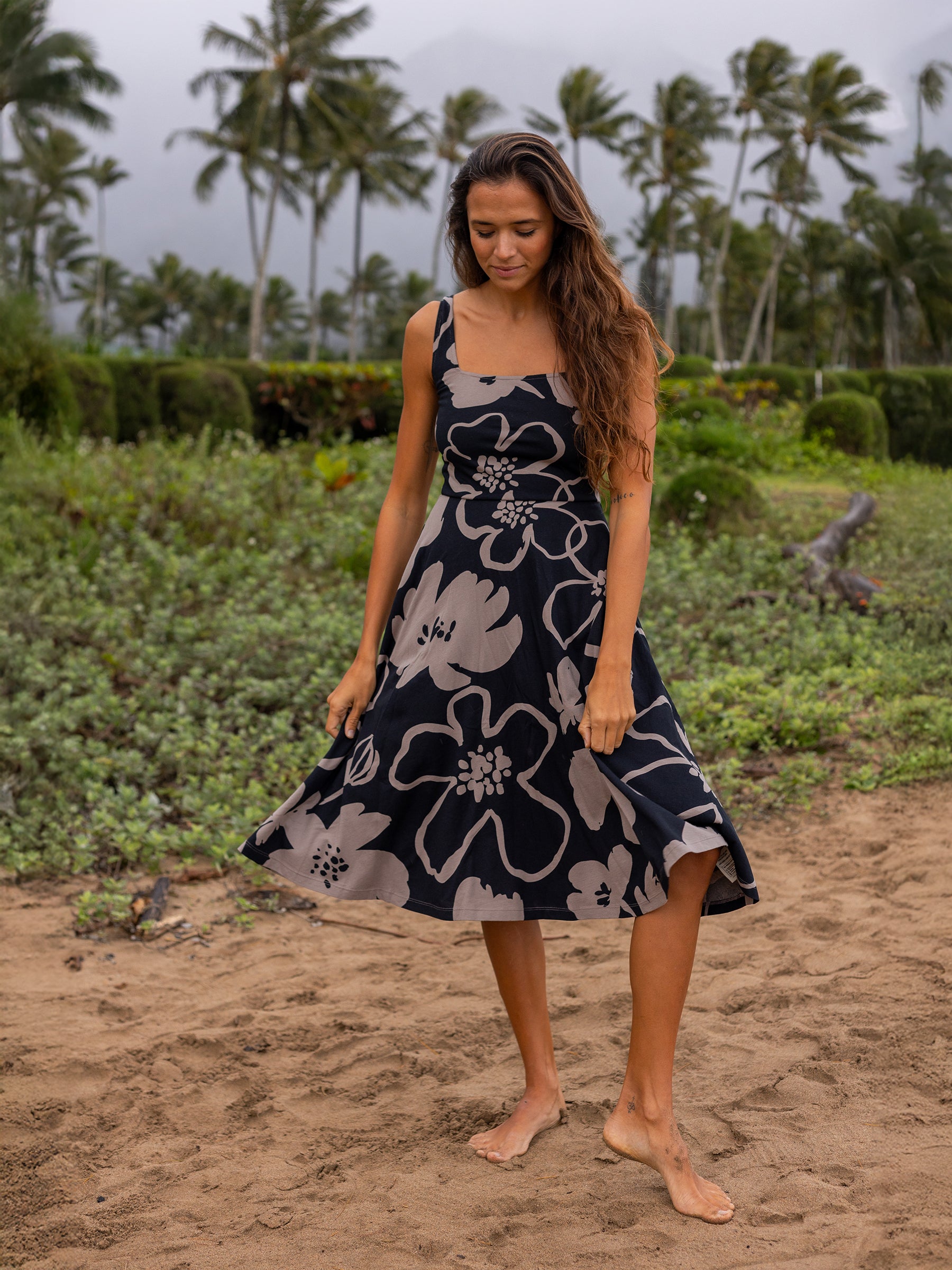 Woman in a Lily Dress, Moonflower print: black organic cotton midi dress with large taupe florals, square neckline, and wide straps, on a beach.
