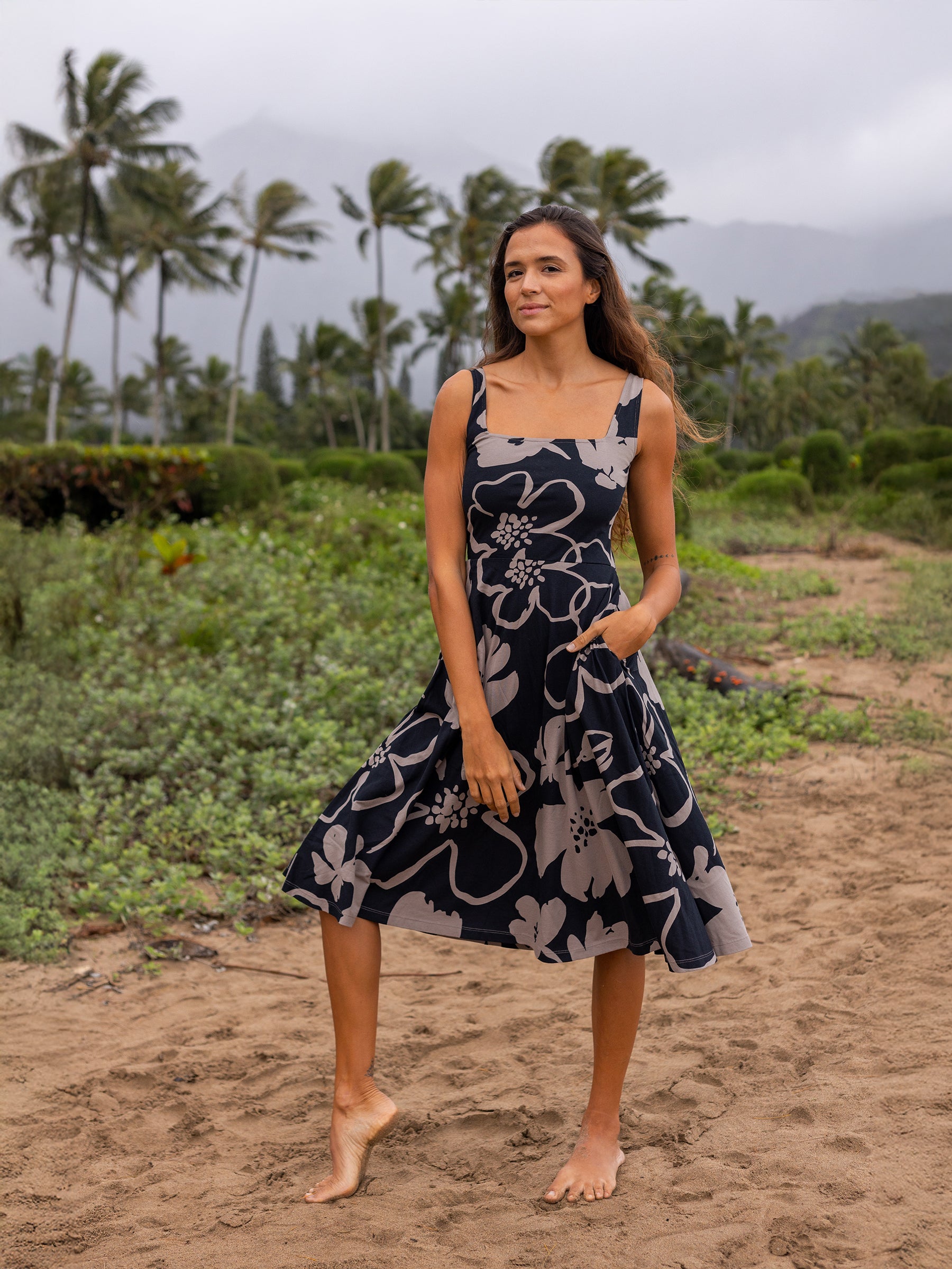 Woman in a black Lily Dress with taupe moonflower print, square neckline, wide straps, and pockets, standing on a beach.