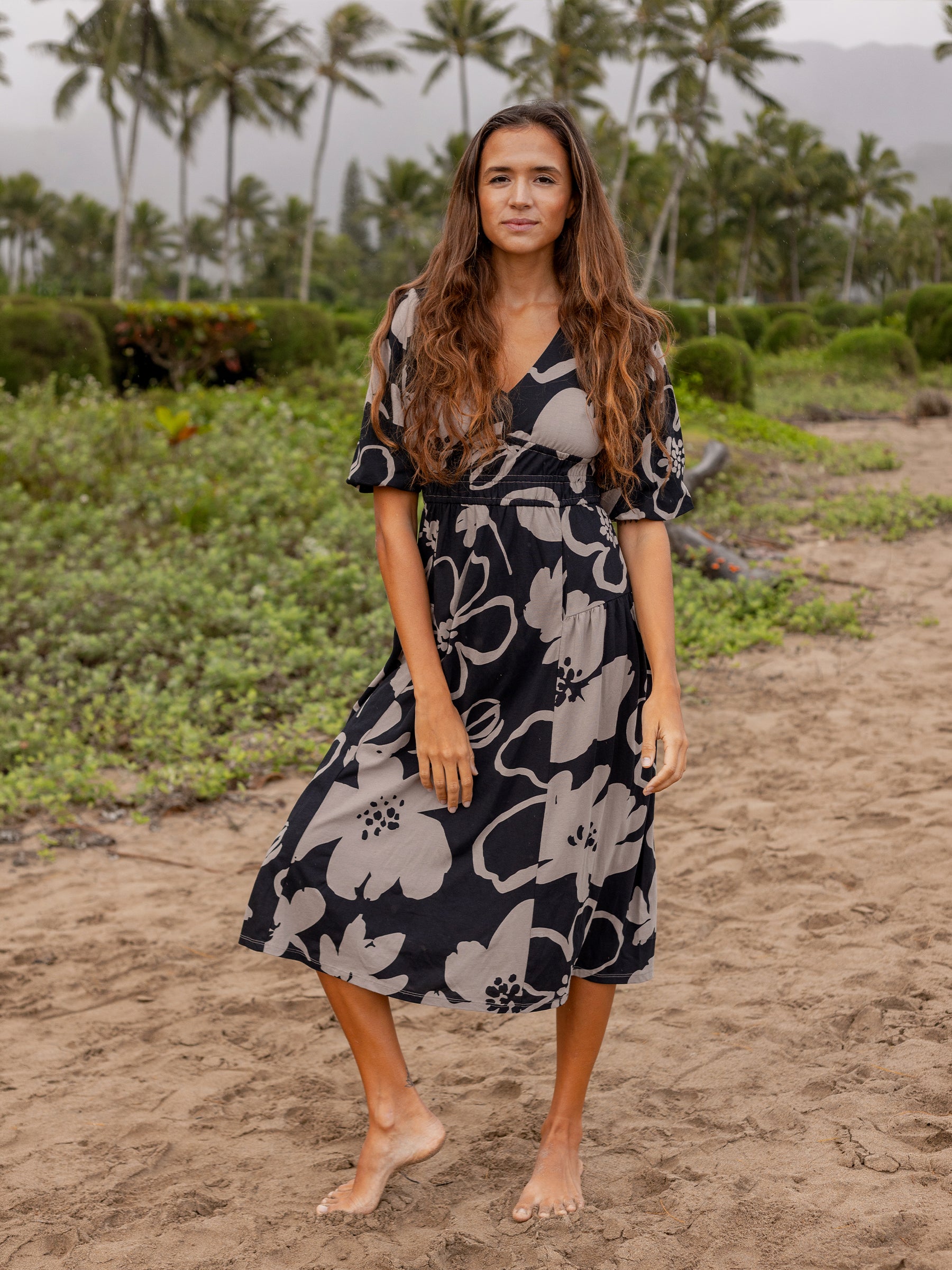 Woman in a black and taupe floral Ophelia midi dress with puffed sleeves and V-neck, standing barefoot on sand.