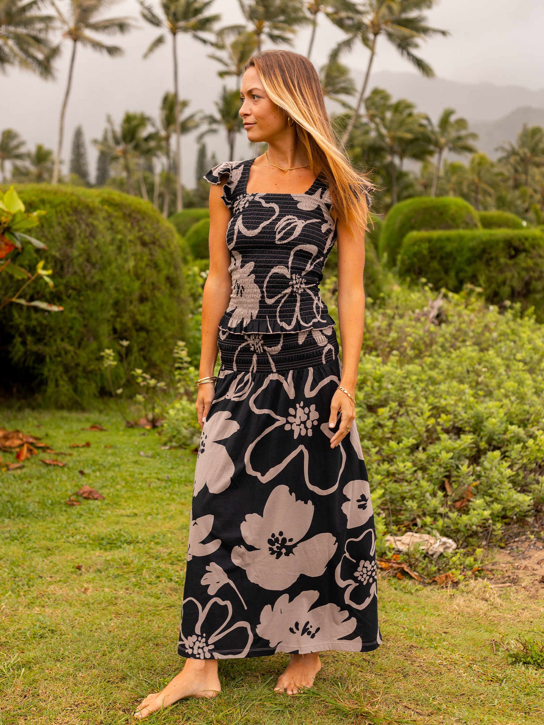 Woman in a black and taupe floral smocked maxi skirt and matching top, standing barefoot outdoors.