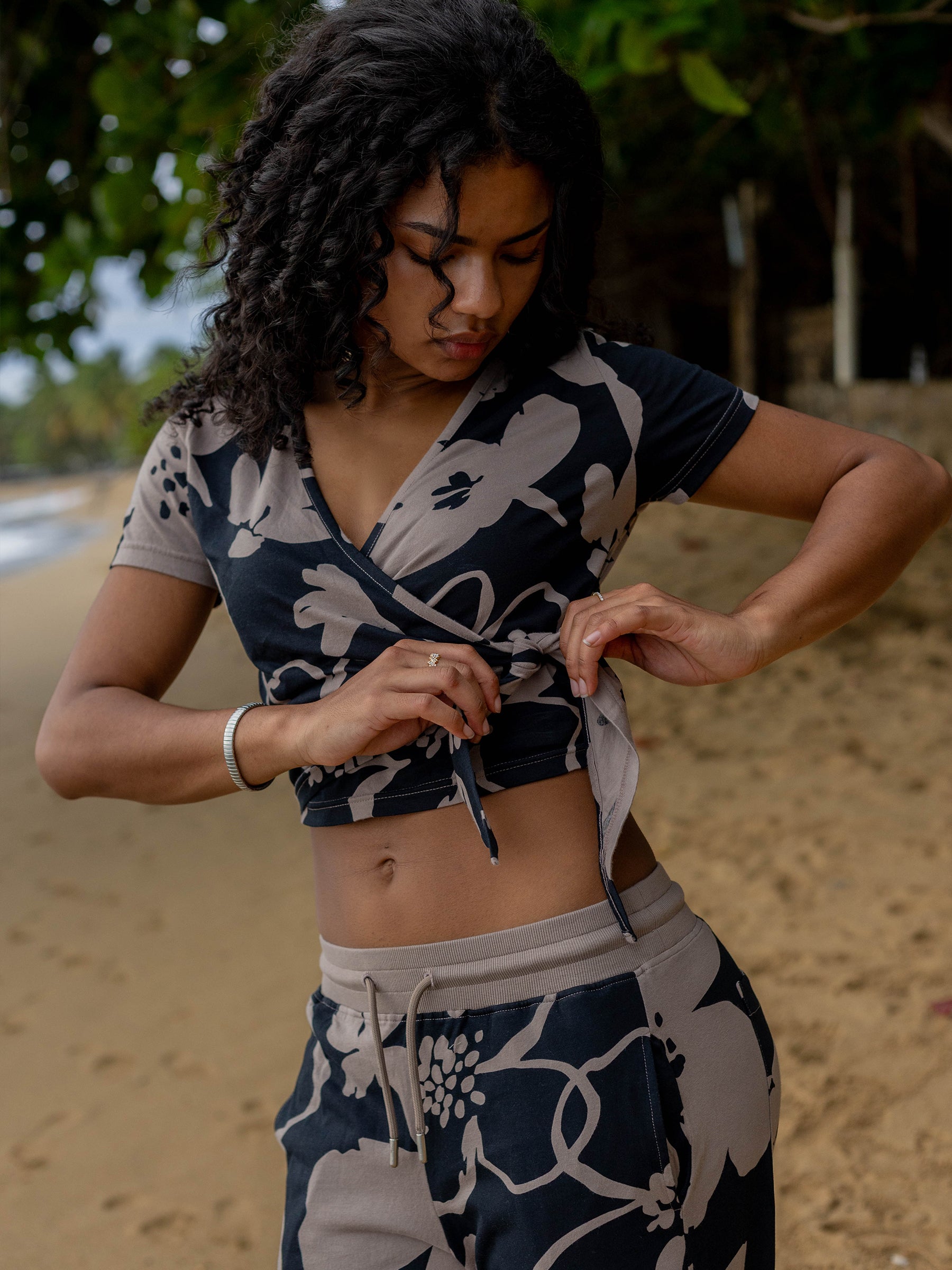 Woman in a Moonflower wrap crop top with large taupe florals, tying the waist on a sandy beach.
