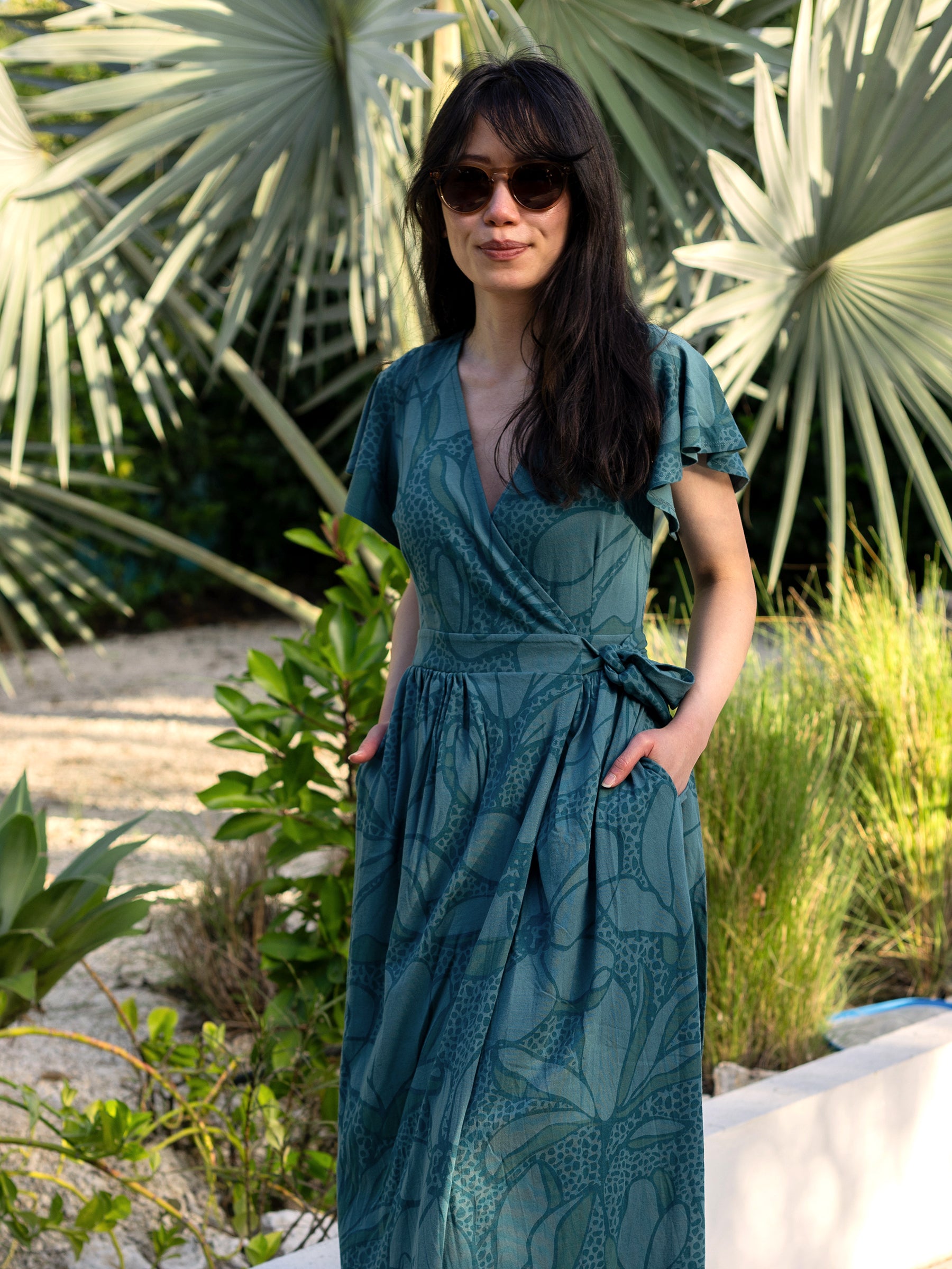 Woman in a teal Isla maxi wrap dress with large florals, flutter sleeves, V-neck, and pockets.
