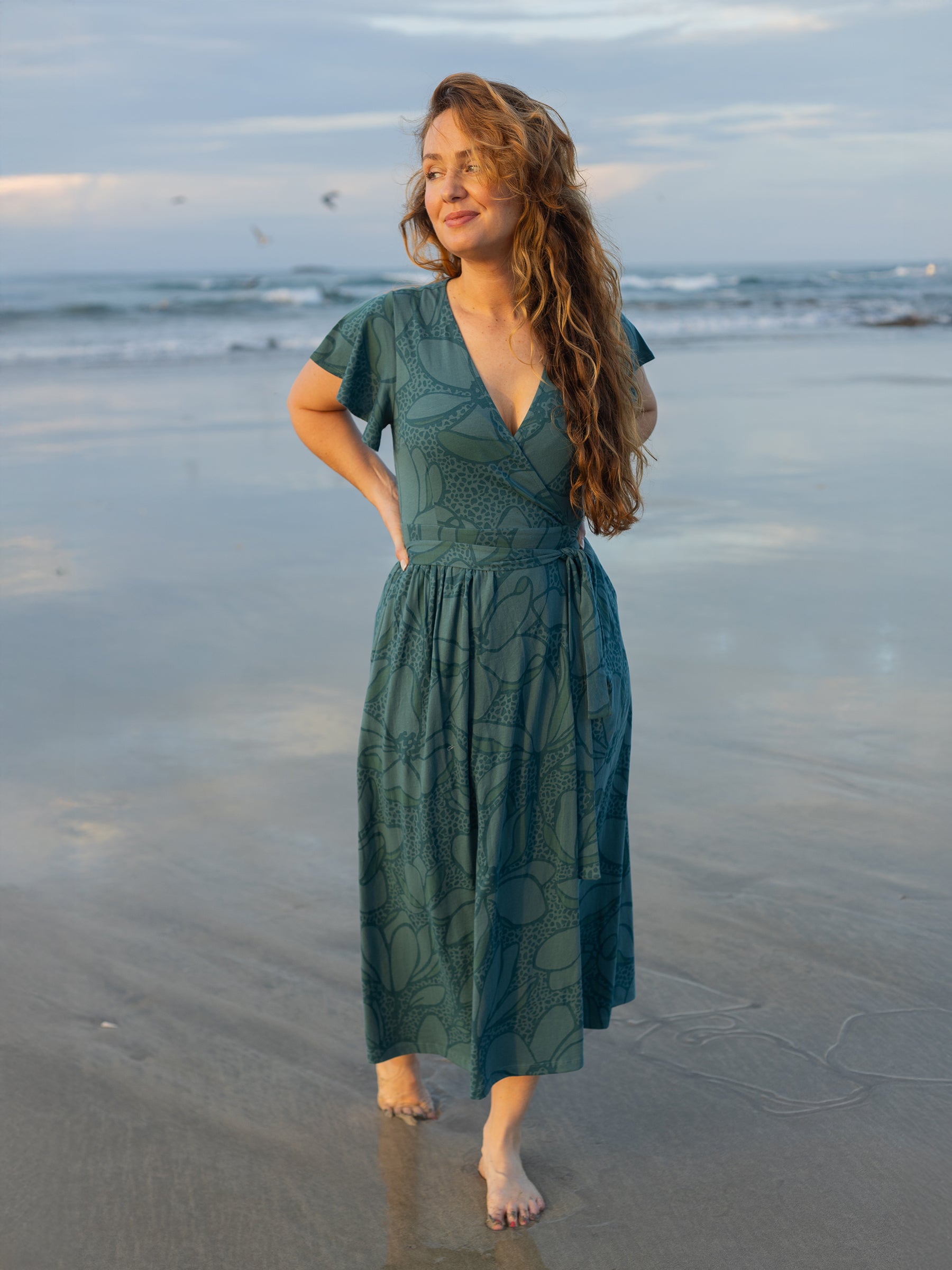 Woman in a seaside teal Isla maxi wrap dress with large florals, flutter sleeves, and V-neck on a beach.