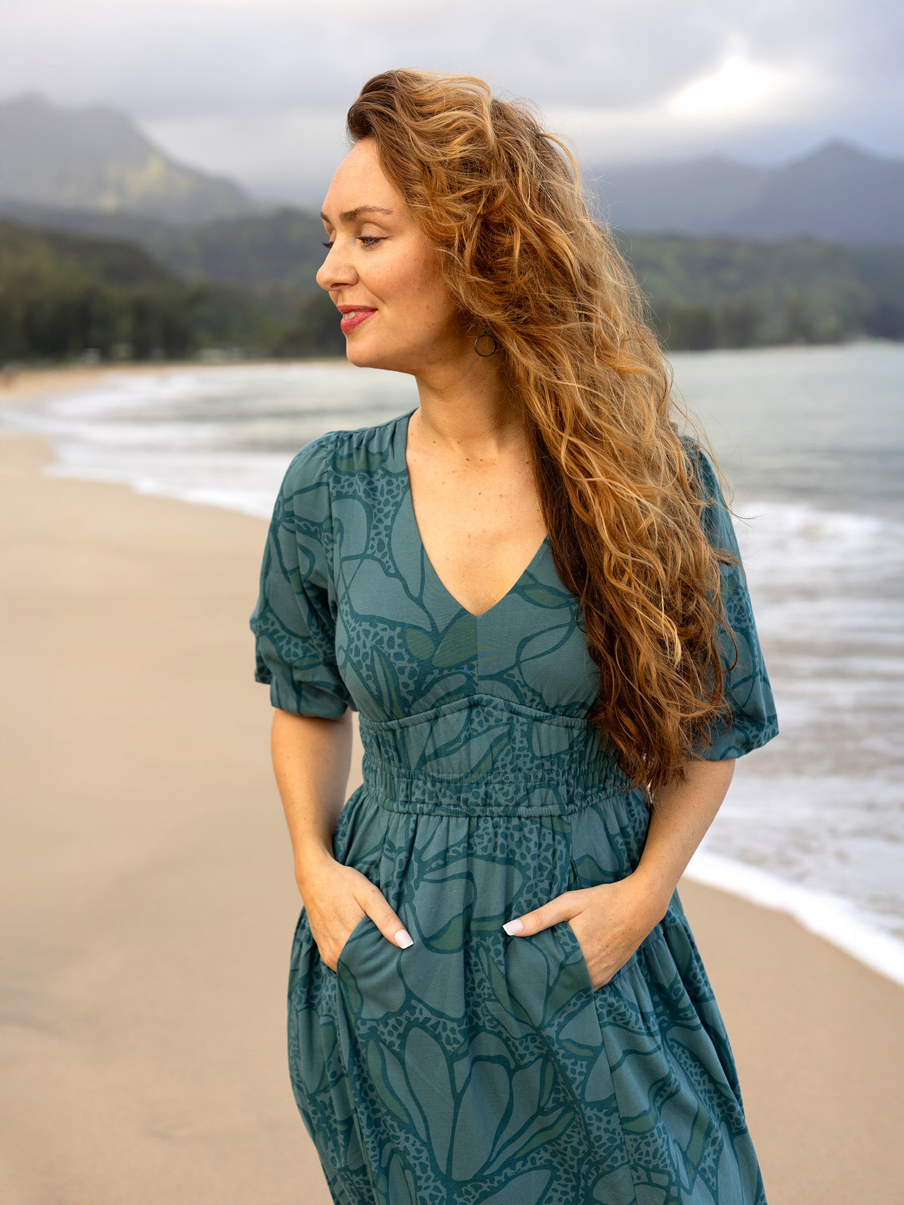 Woman in a teal organic cotton Ophelia Dress with large florals, V-neck, puffed sleeves, and smocked waist, on a beach.