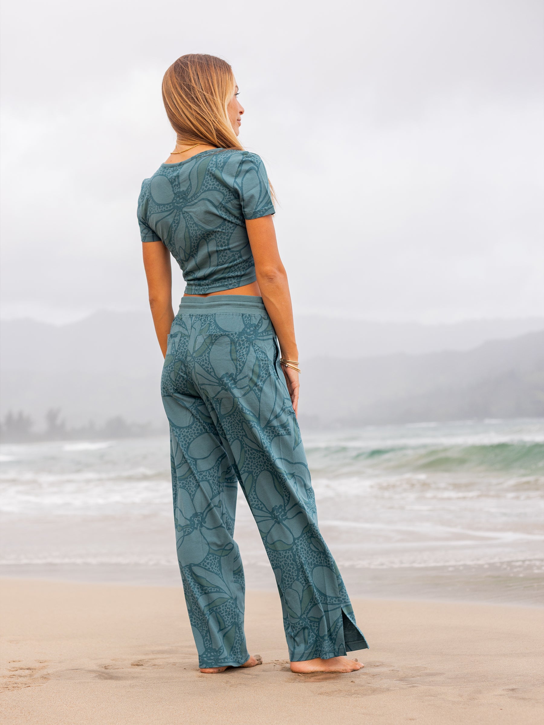Woman in a seaside teal organic cotton wrap crop top and matching wide-leg pants with floral print on a beach.