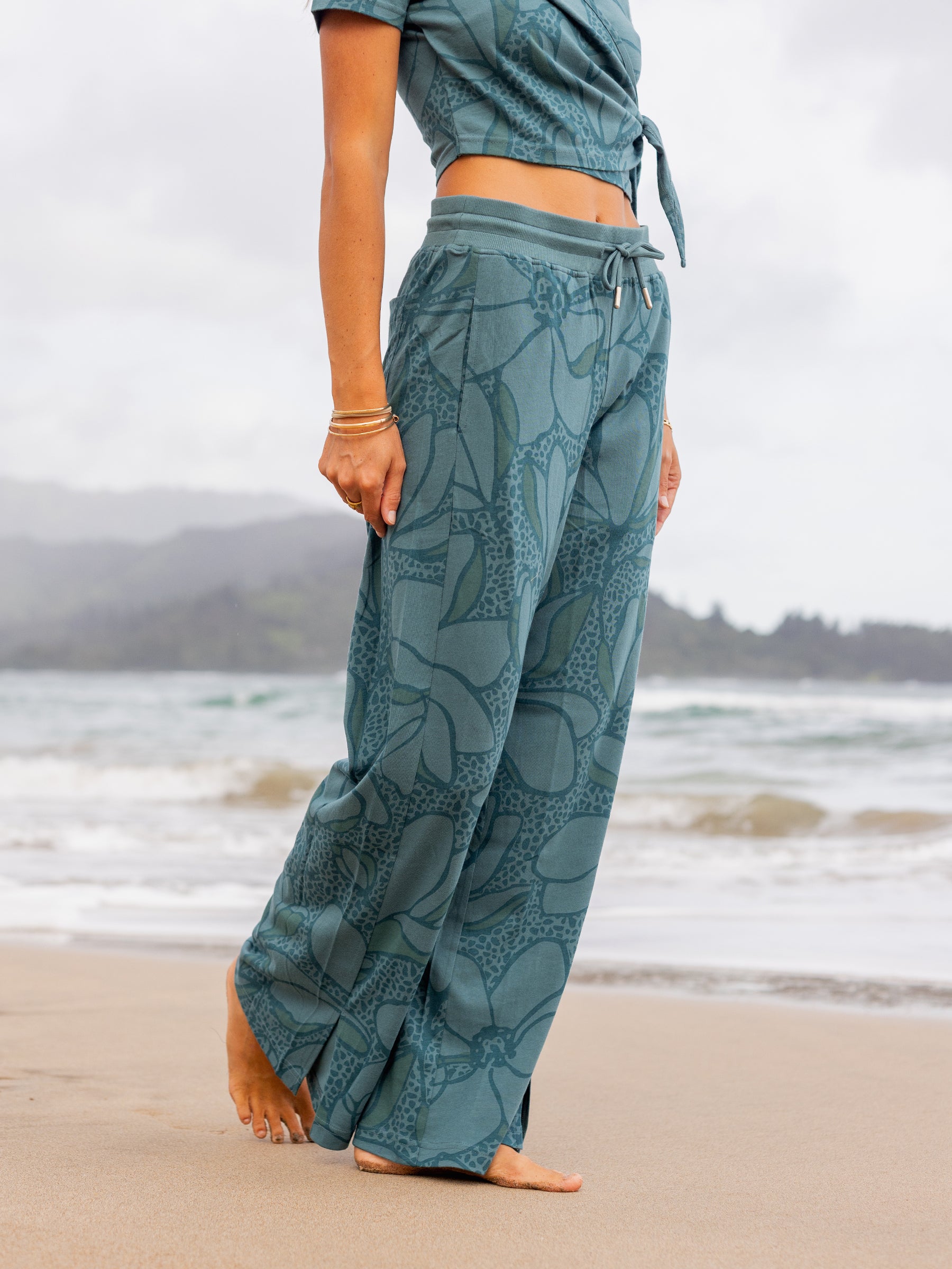 Teal organic cotton wide-leg pants with large floral print, side slits, and drawstring waist on a beach.