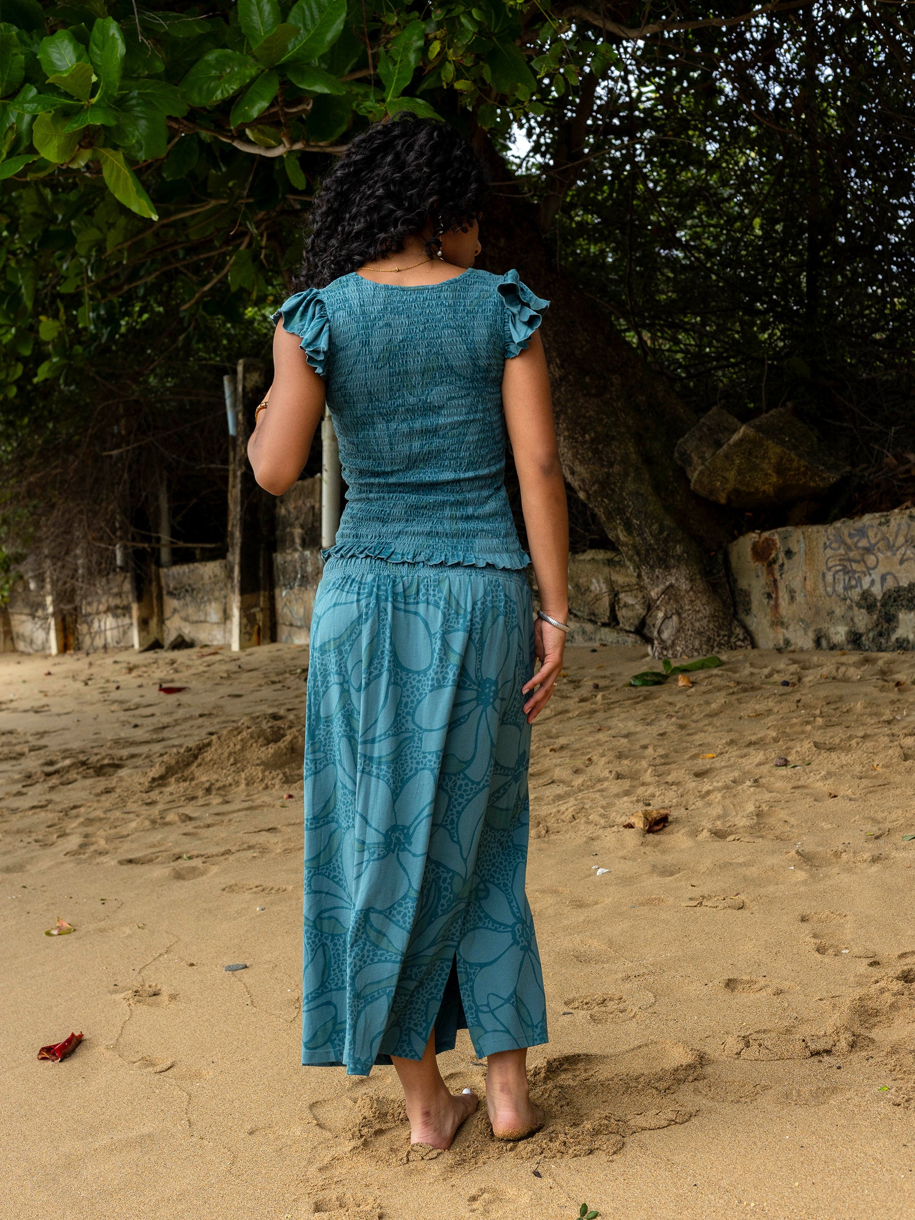 Woman in a teal smocked top and matching maxi skirt with a side slit and floral pattern on a sandy beach.