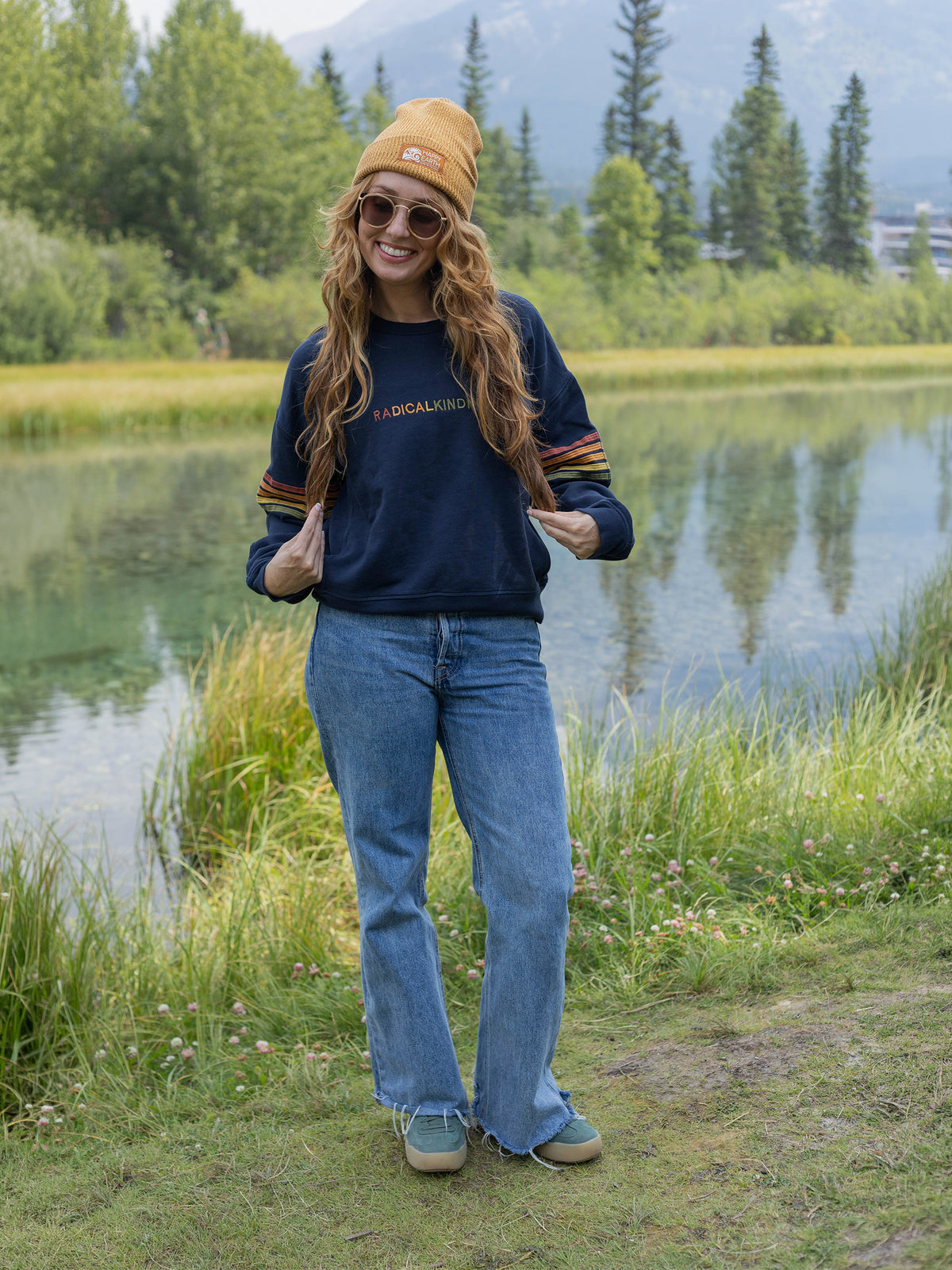 Model wearing the Radical Kindness organic cotton sweatshirt in navy with jeans and a beanie by a mountain lake