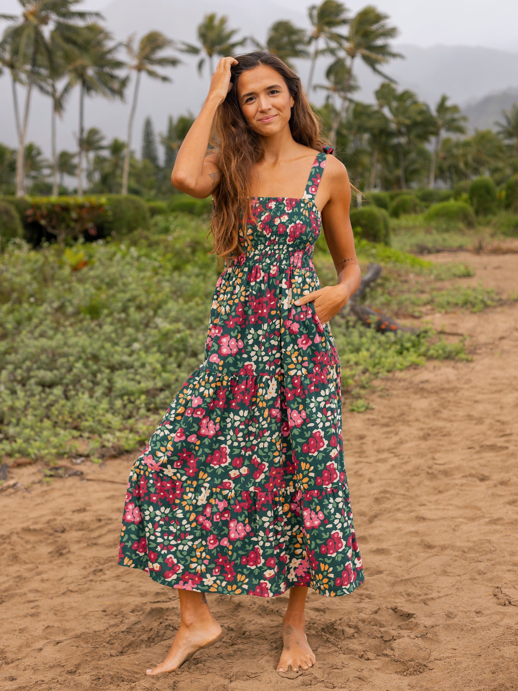 Woman in a green Evie Smocked Dress with red and pink wildflowers, smocked bodice, and tiered maxi skirt on a beach.