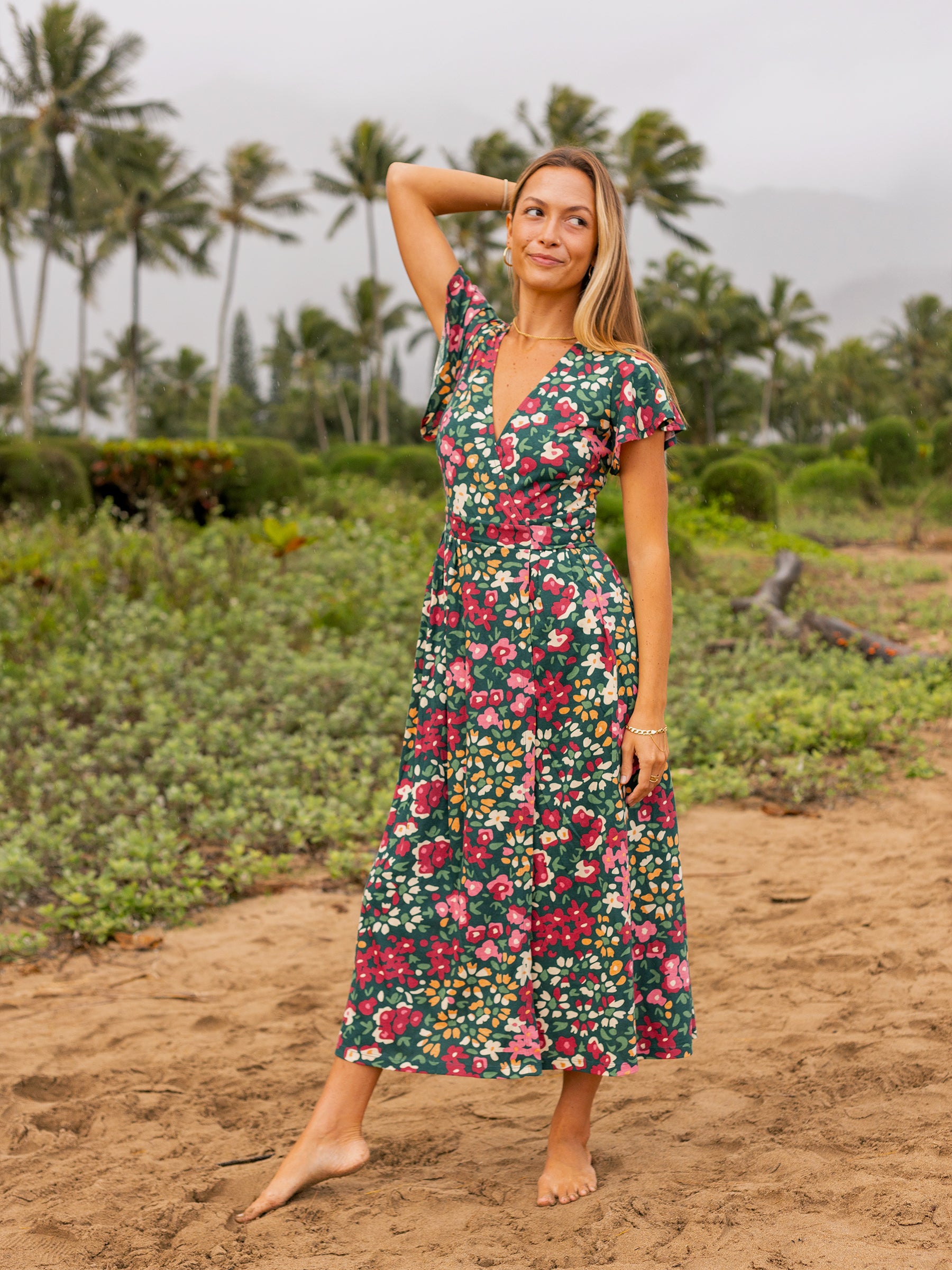 Woman in a green Isla maxi wrap dress with red and pink wildflowers, flutter sleeves, and V-neck, on a beach.