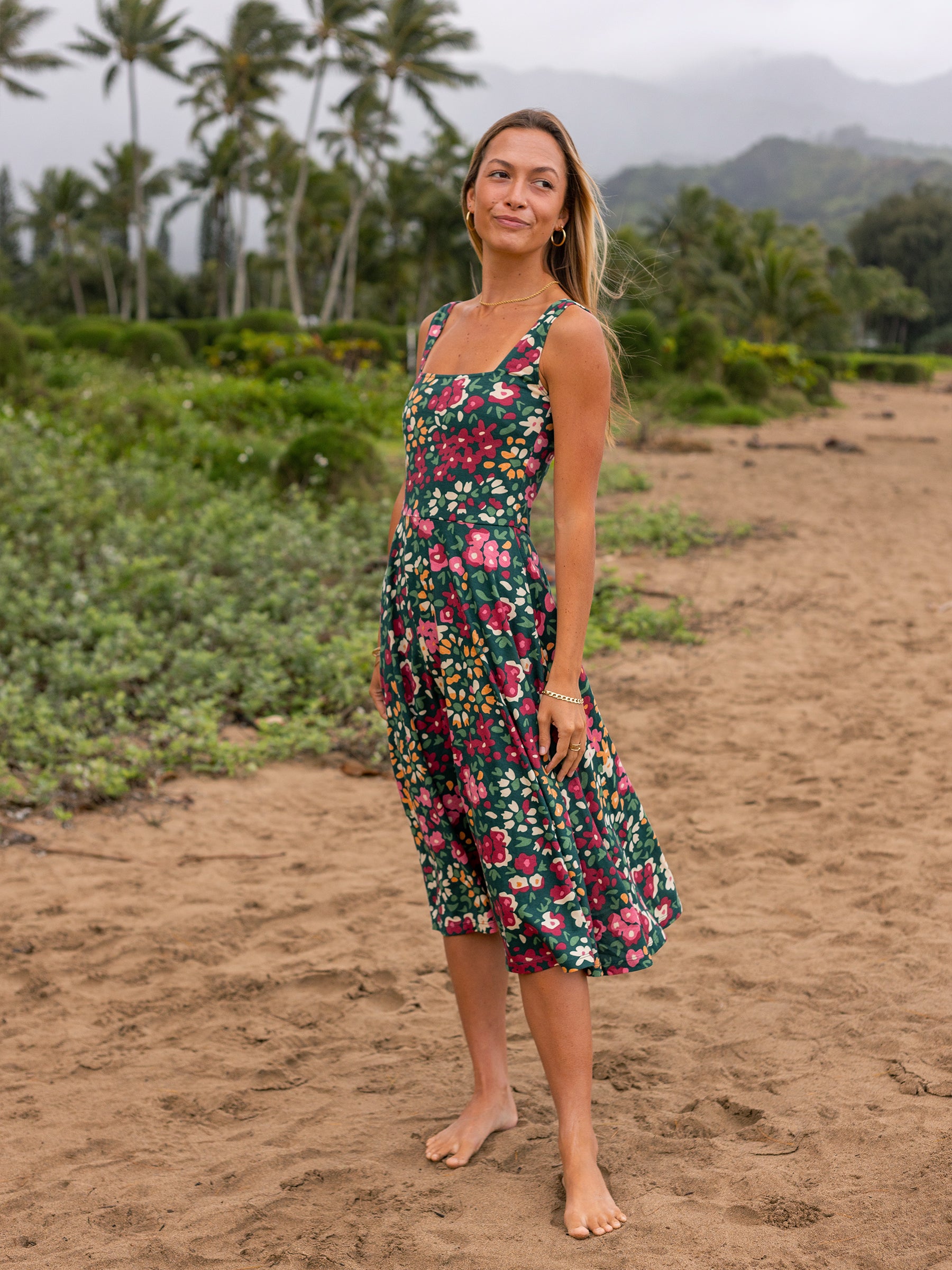 Woman in a green Lily Dress with colorful wildflowers, square neckline, and midi skirt on a sandy beach.