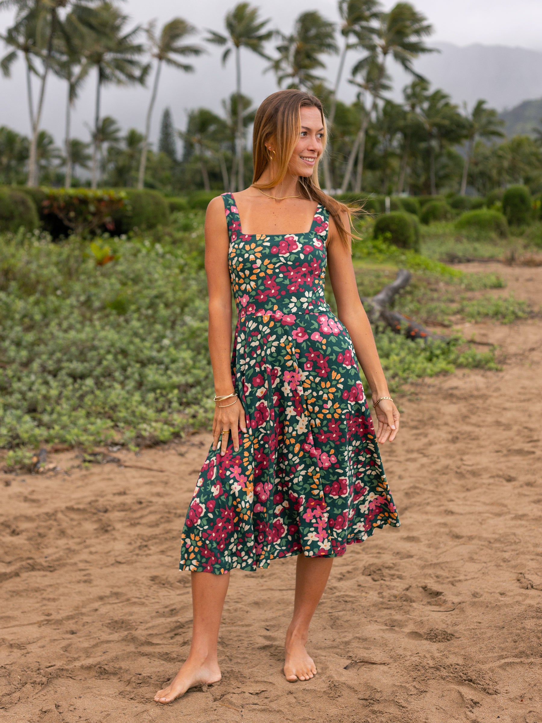 Woman in a green Lily Dress with a colorful wildflower print, square neckline, and flowy midi skirt on a beach.