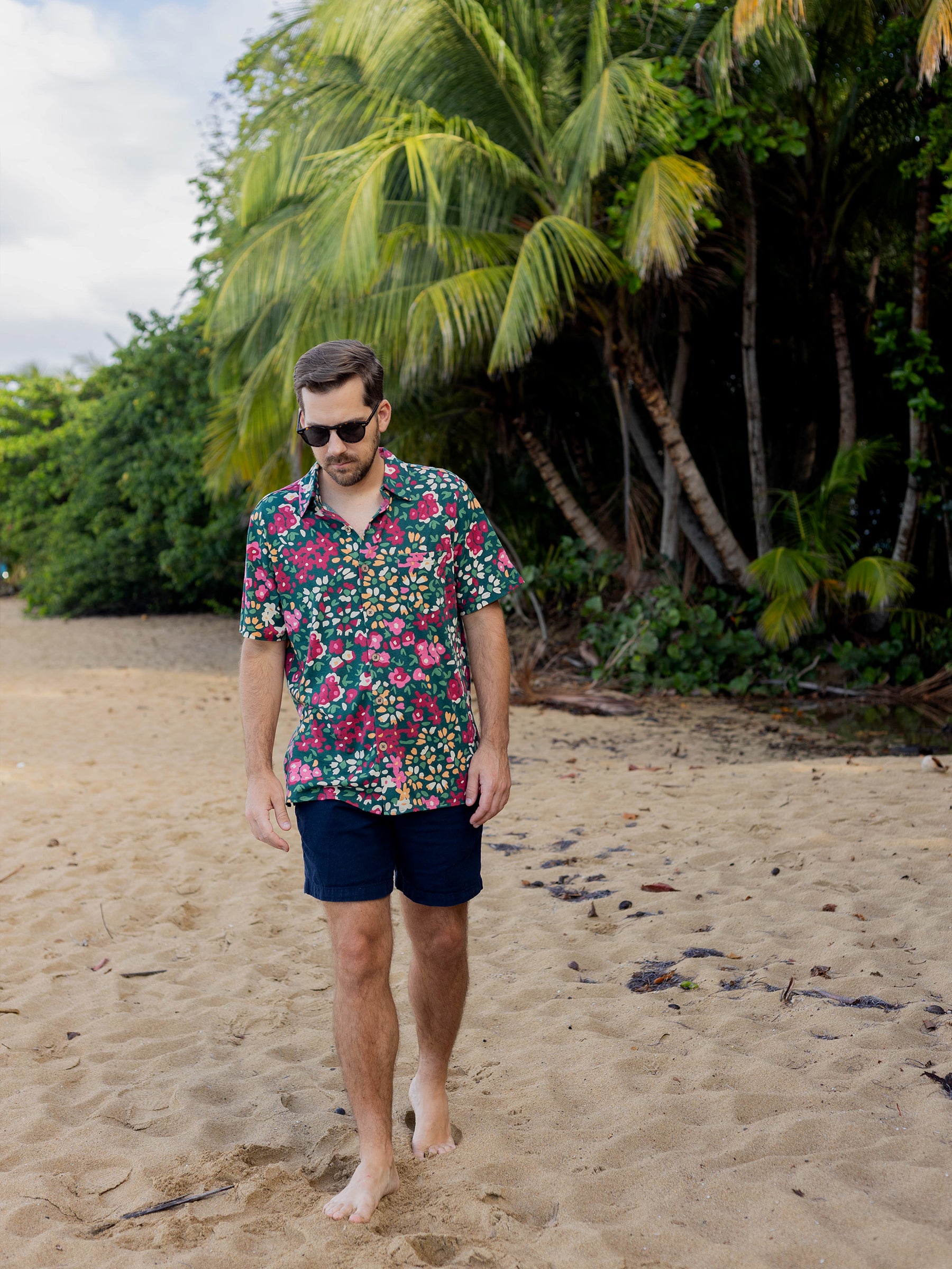 Man in a green Rincon Shirt with colorful wildflowers, dark shorts, and sunglasses walking barefoot on a sandy beach.