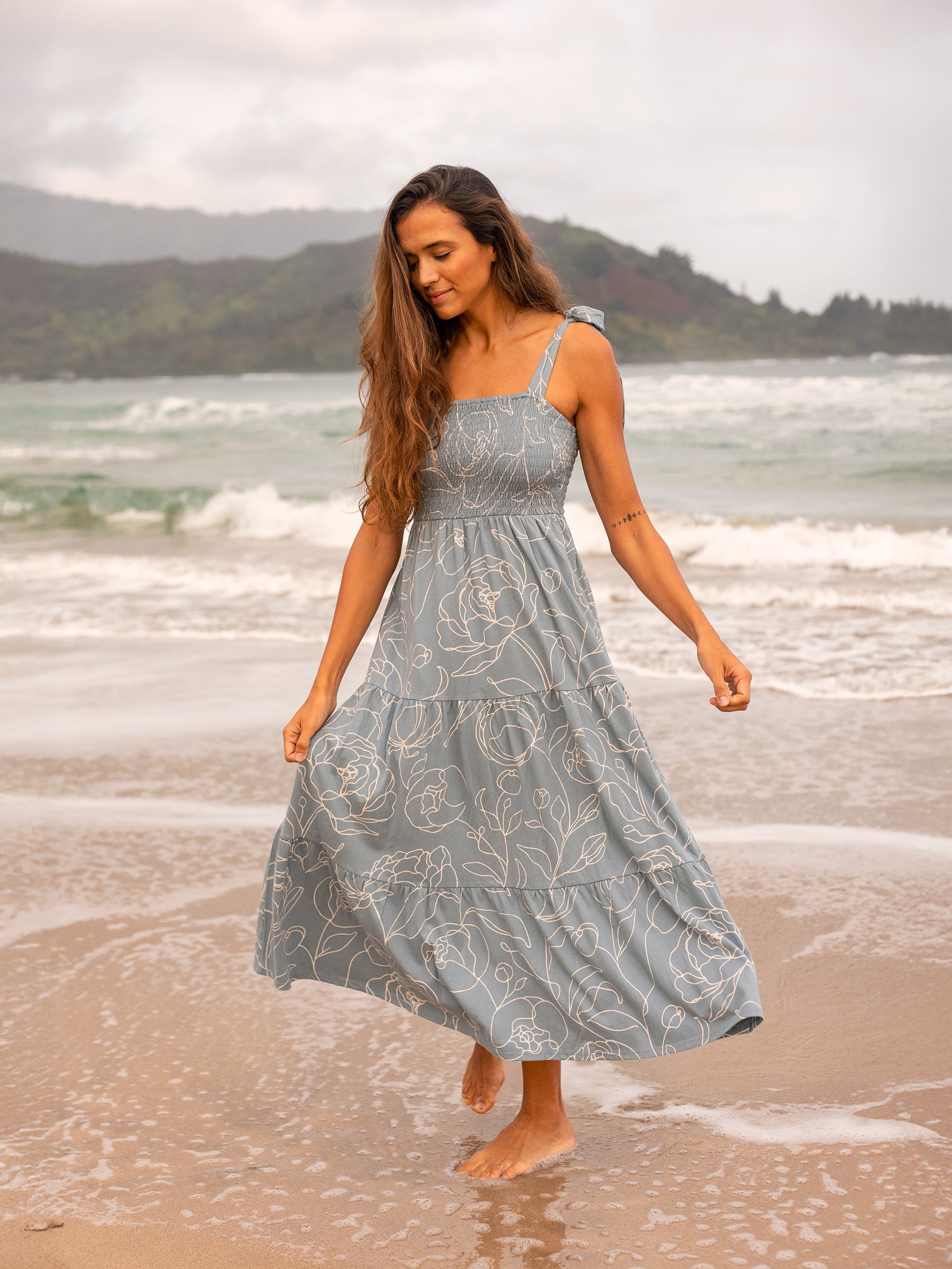 Evie Smocked Dress in Garden Rose: a woman in a light blue tiered maxi dress with white floral print on a beach.