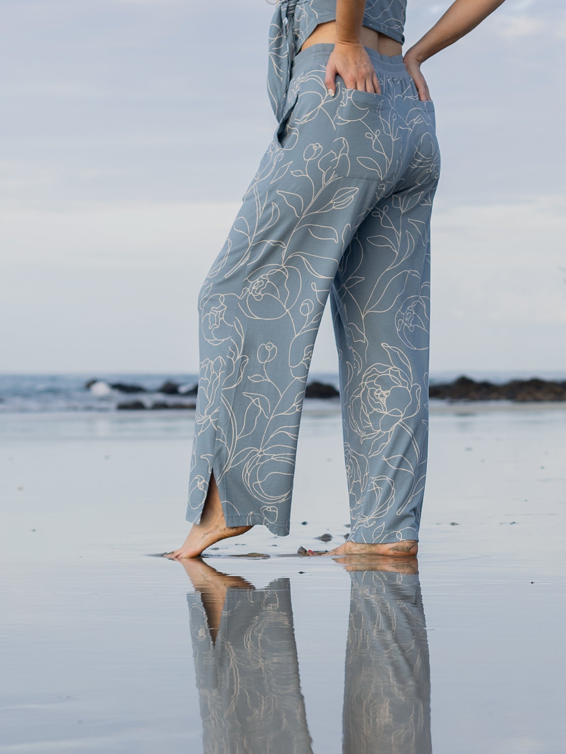 Woman in blue wide-leg organic cotton pants with white hand-drawn rose print and side slits, standing on a beach.