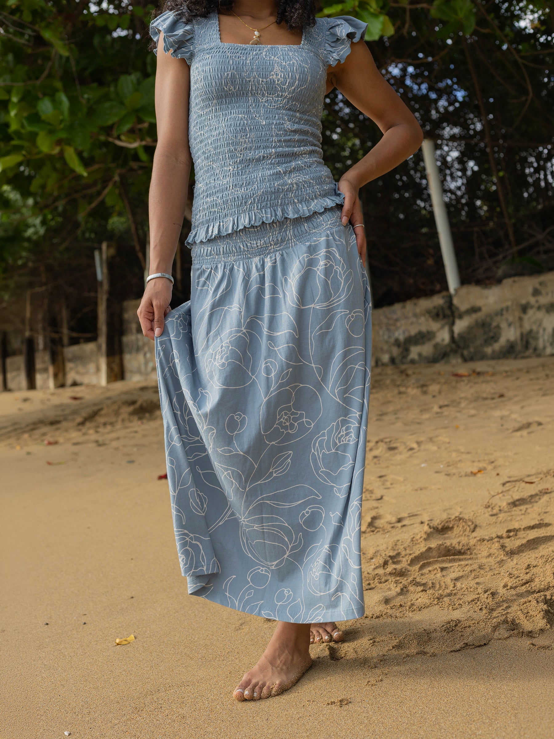 Woman in a blue organic cotton smocked maxi skirt with white hand-drawn roses and a side slit on a beach.