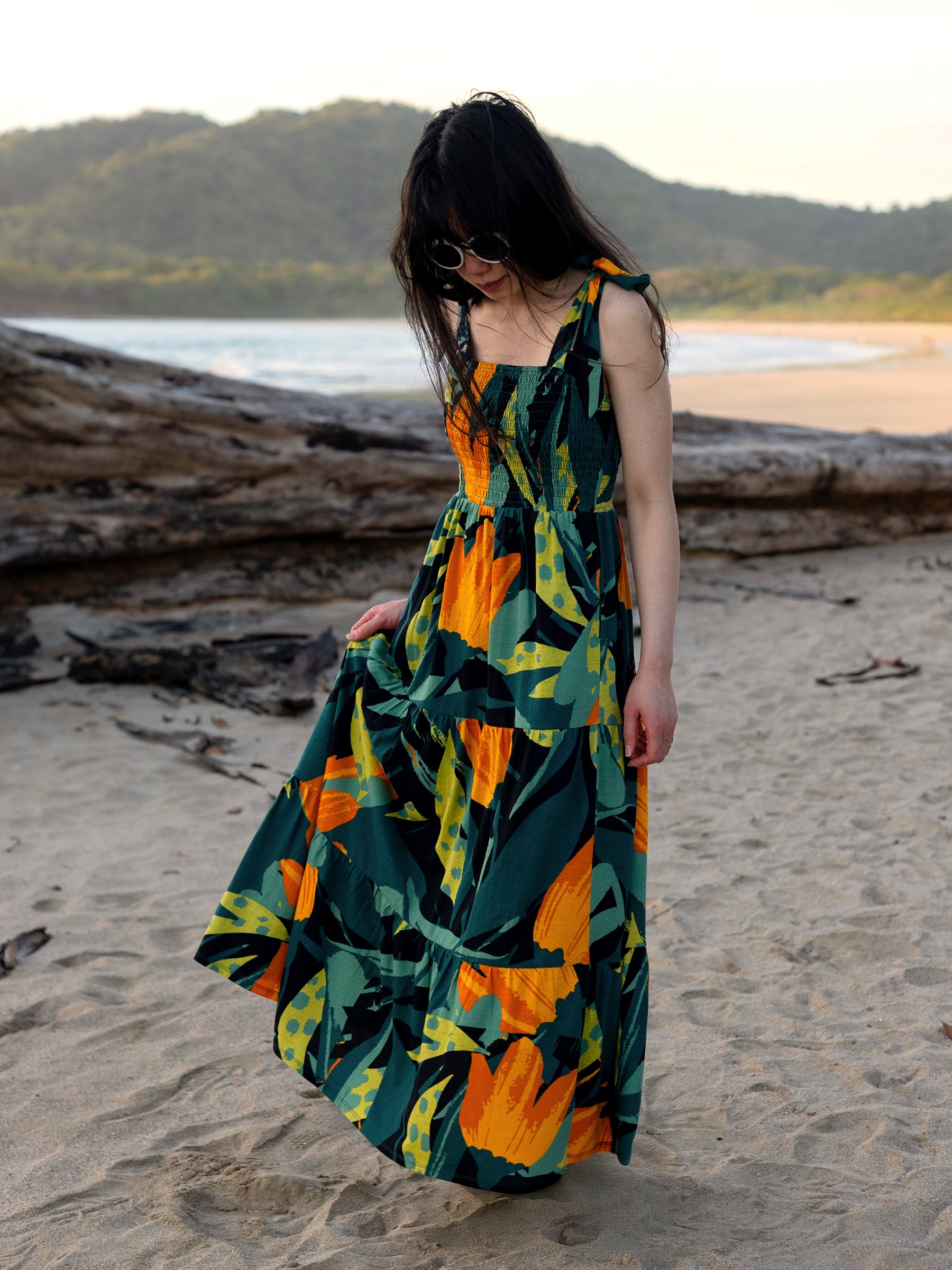 Woman in a green, orange, and yellow floral Evie Smocked Maxi Dress with ribbon ties on a sandy beach.
