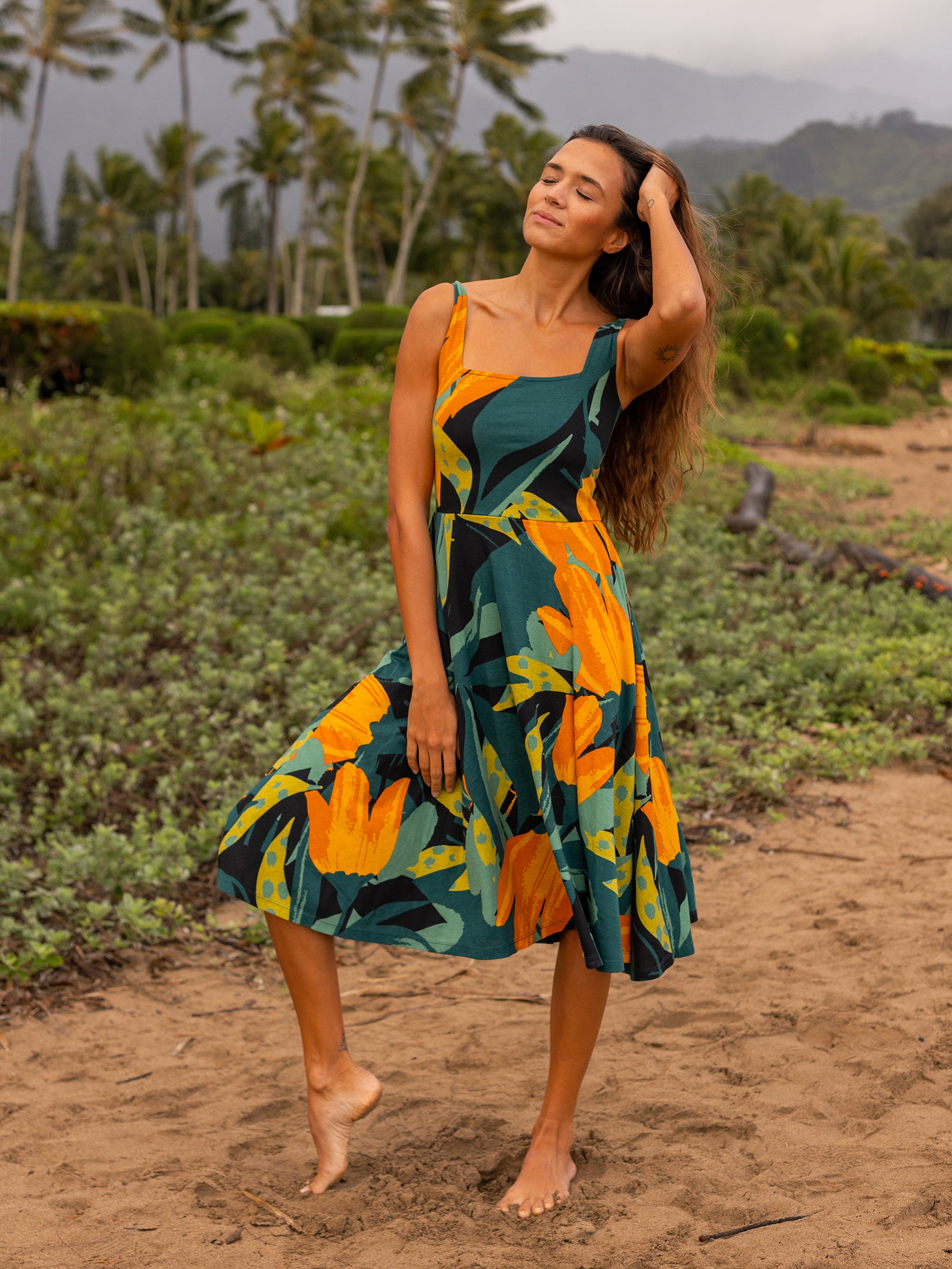 Woman in a Hanalei Lily Dress: green organic cotton midi with orange floral print, square neckline, and wide straps.