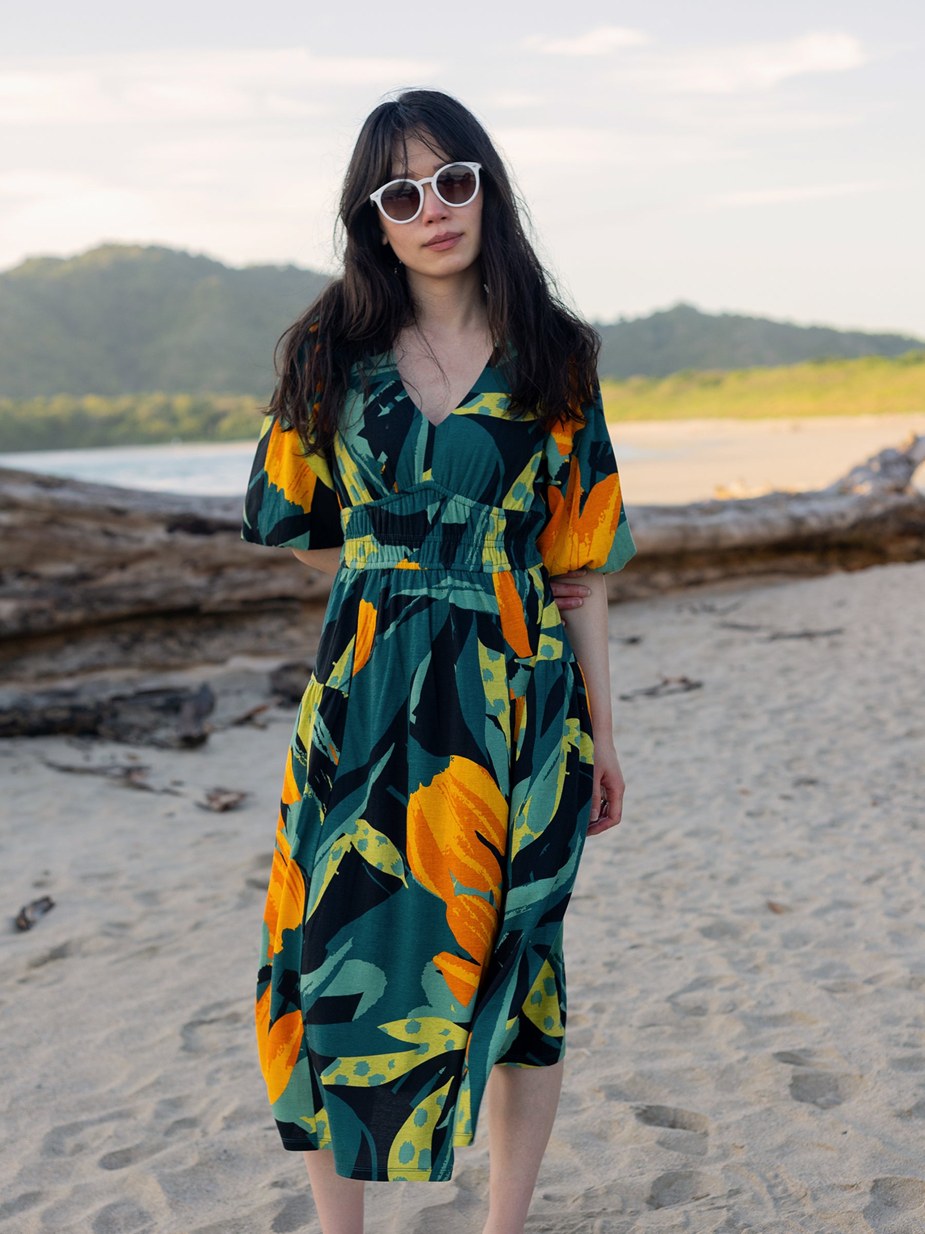 Ophelia Dress: Woman on beach wearing a green, yellow, and orange floral midi dress with V-neck and puffed sleeves.