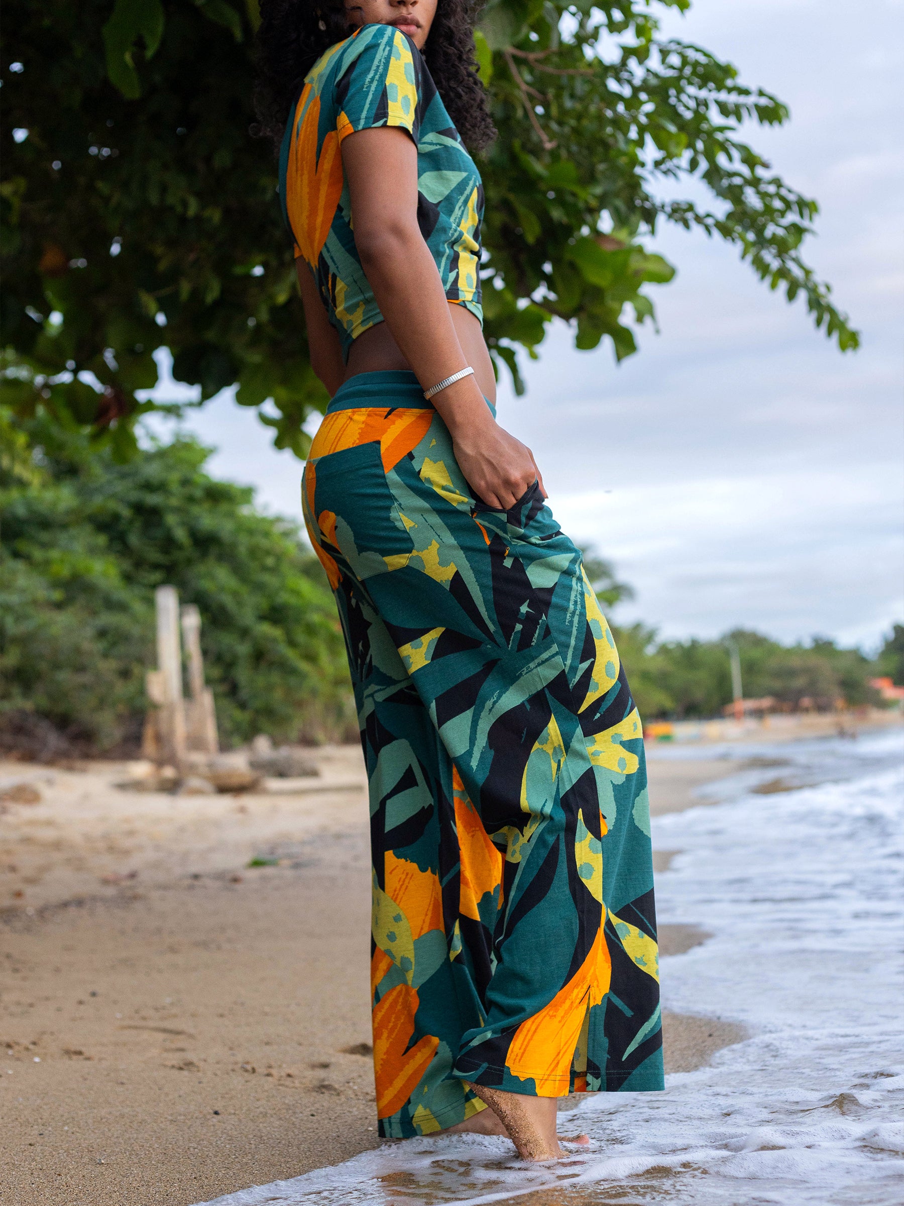 Woman in Hanalei wide leg pants and matching top with green, yellow, and black tropical print on a beach.
