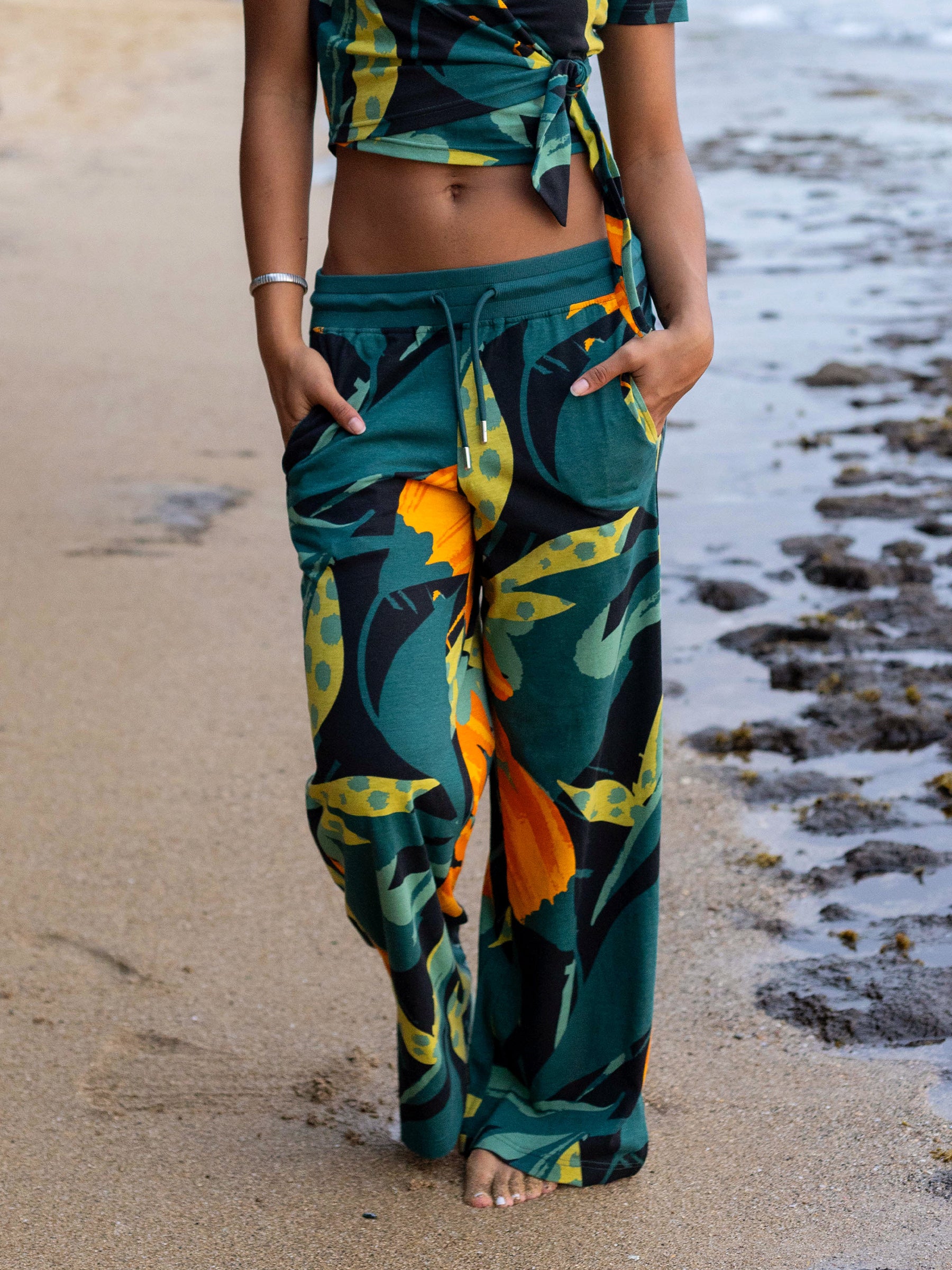 Woman wearing Hanalei wide-leg organic cotton pants with a green, black, and orange tropical print, walking on a sandy beach.
