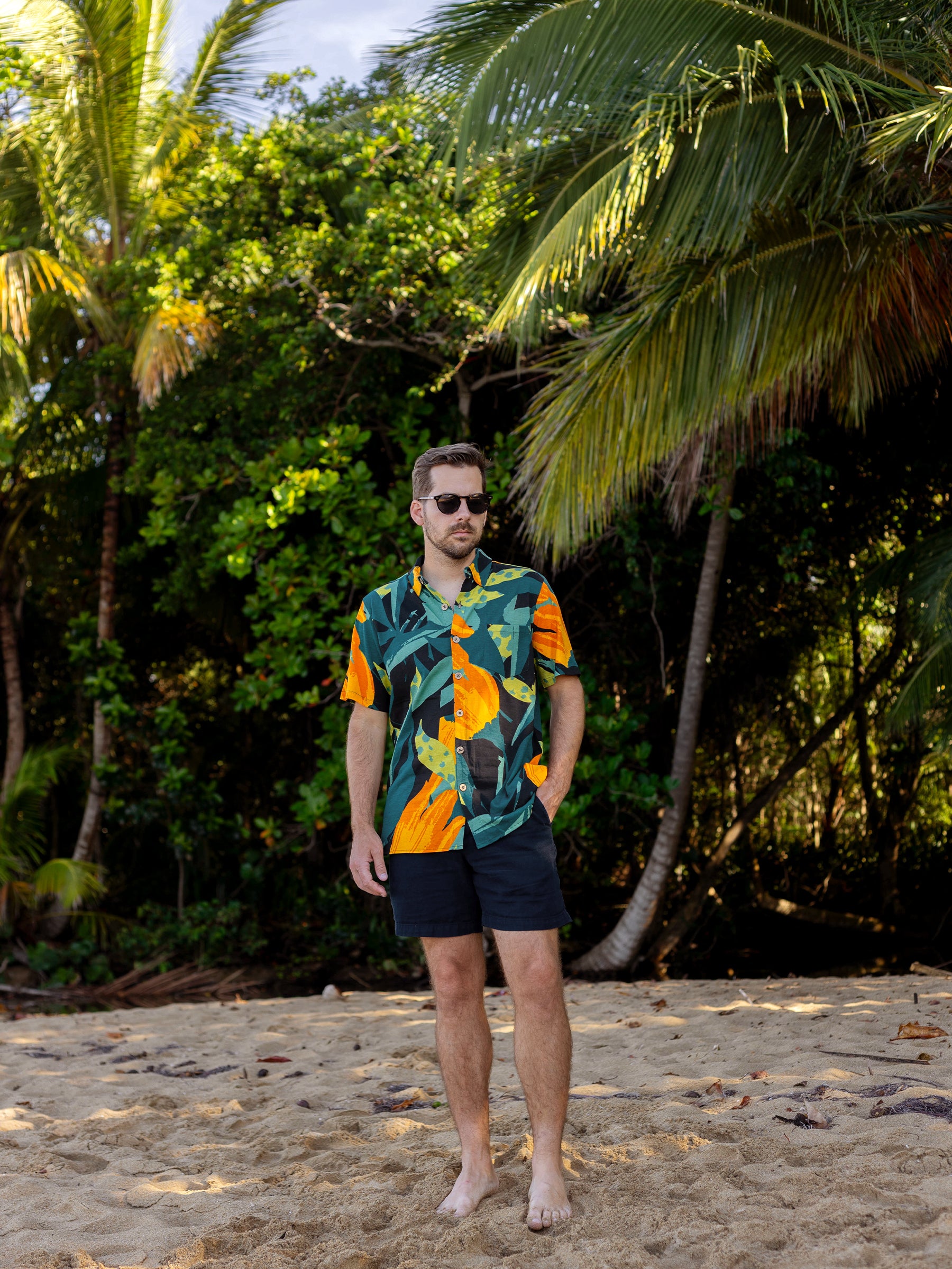 Man in Rincon Shirt Hanalei, a green and orange floral organic cotton button-up, standing on a sandy beach.