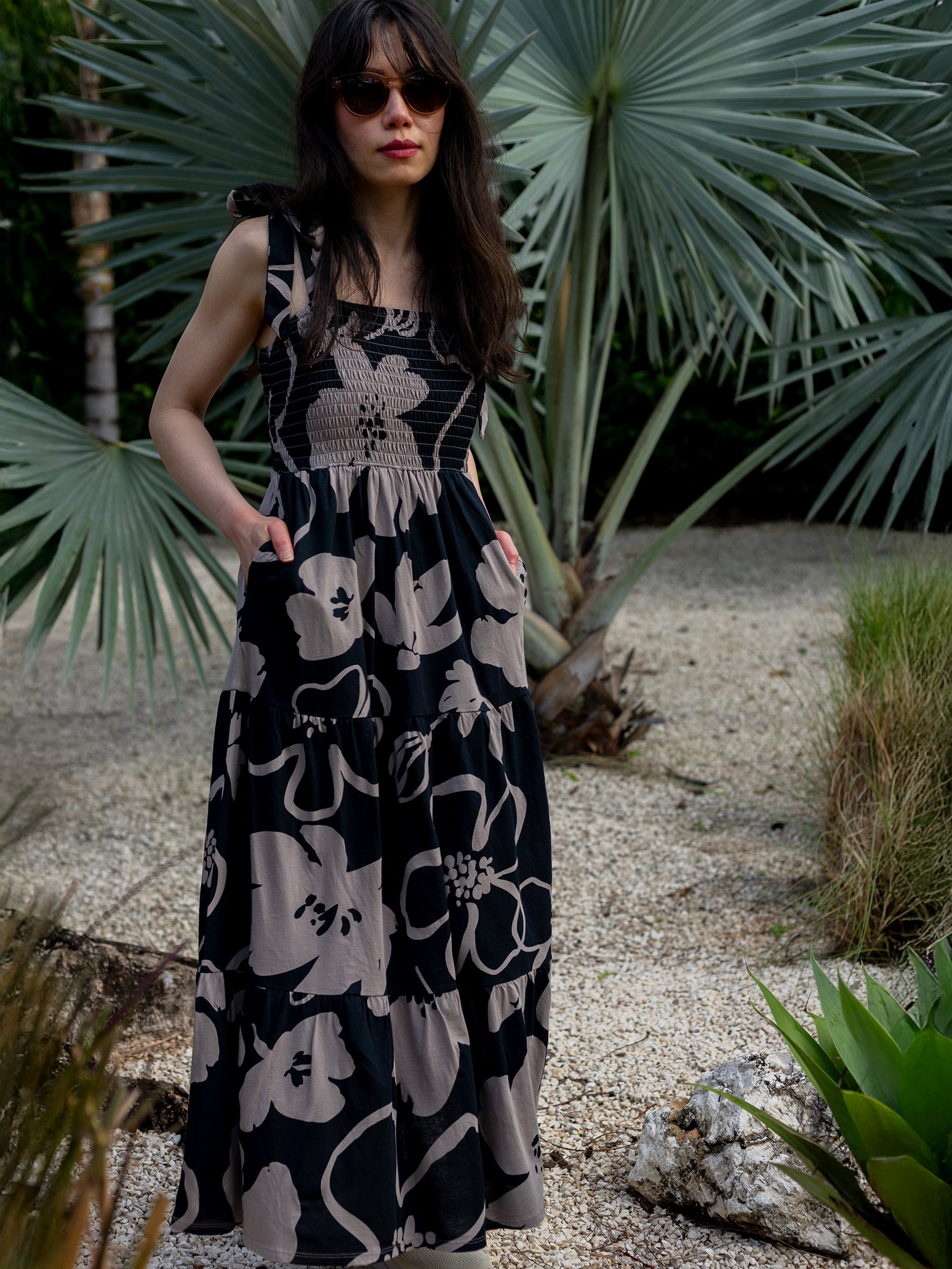 Woman in Evie Moonflower smocked maxi dress with taupe florals, ribbon ties, and pockets, standing in a garden.