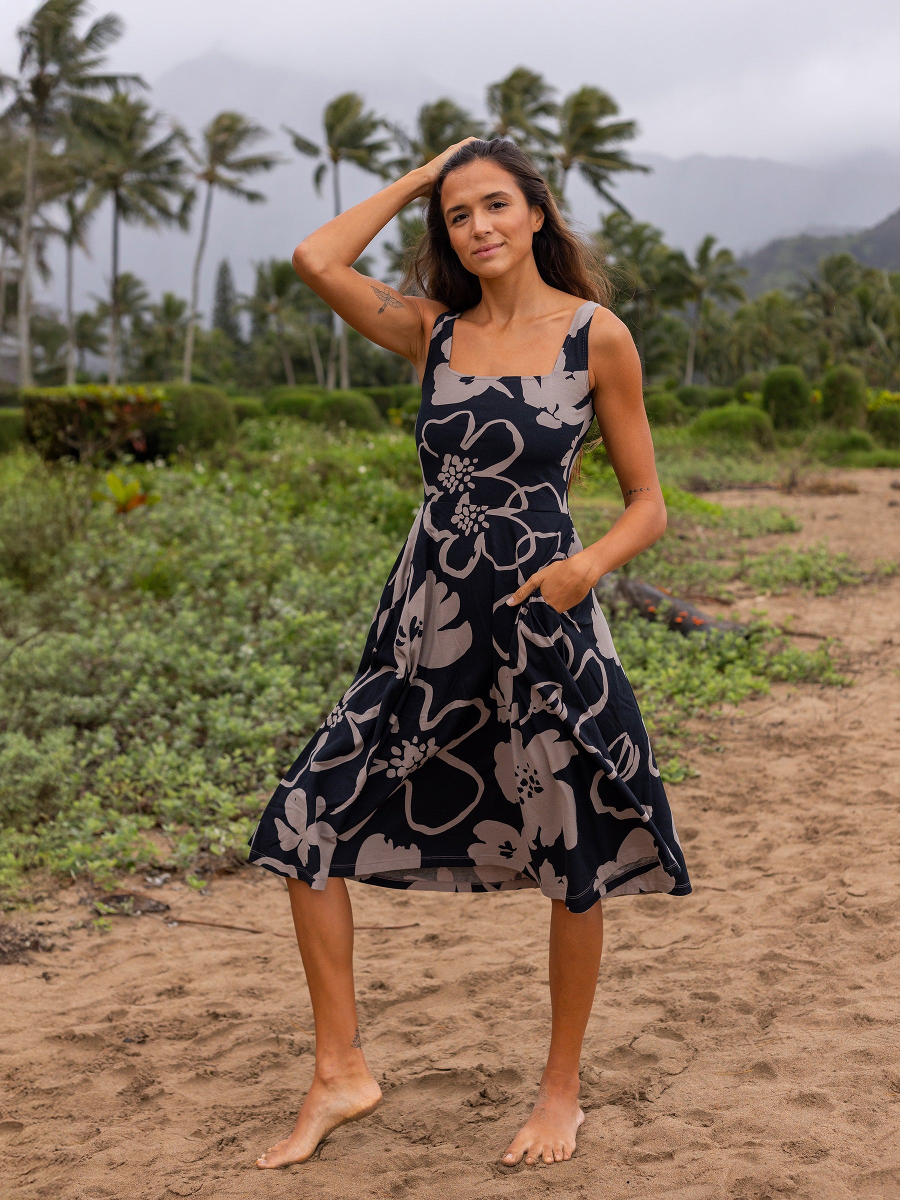 Woman in a black and taupe Moonflower Lily Dress with large floral print, square neckline, and pockets, standing on a beach.