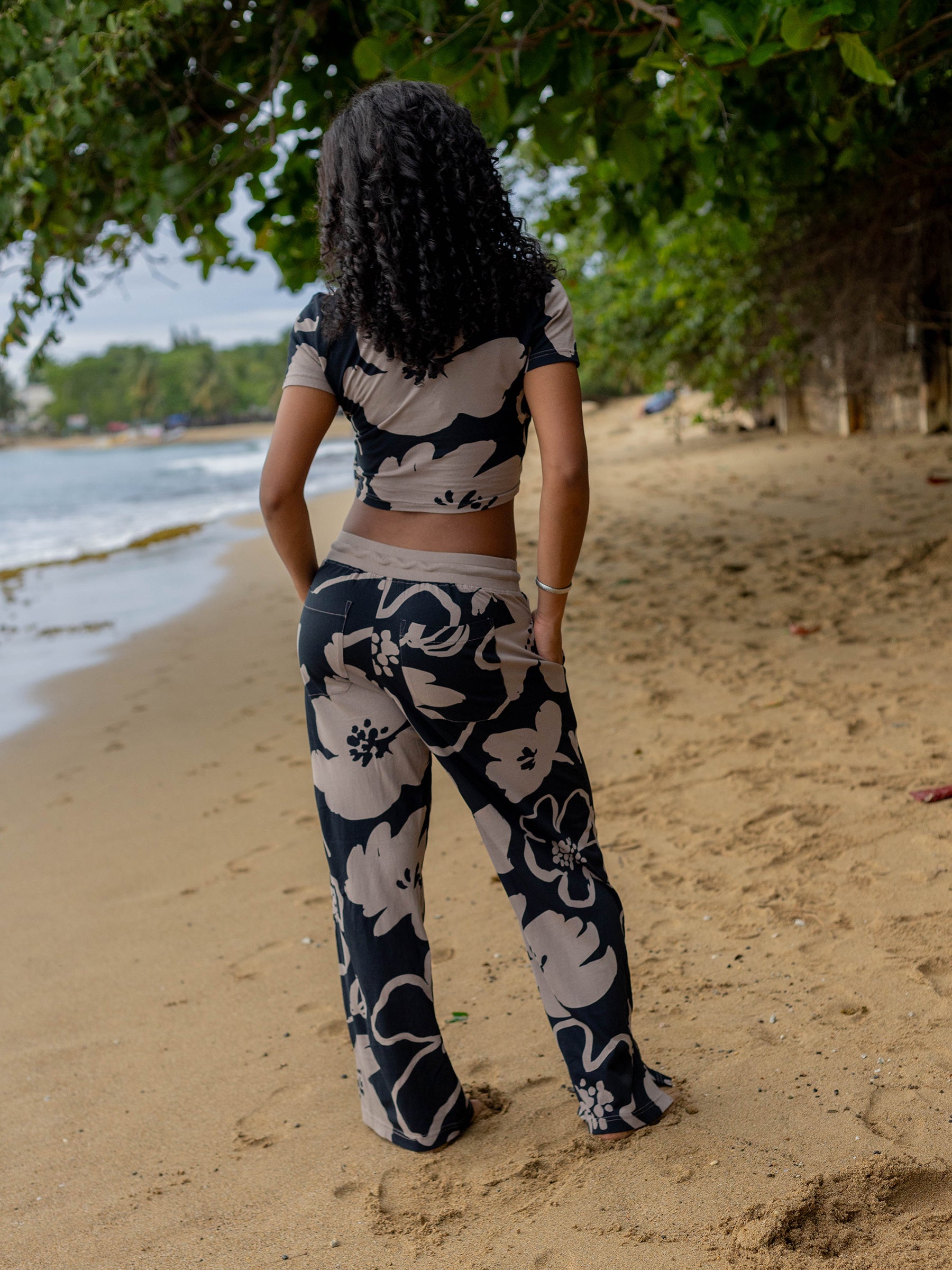 Woman in Moonflower wrap crop top and matching pants with large taupe florals on black, standing on a sandy beach.