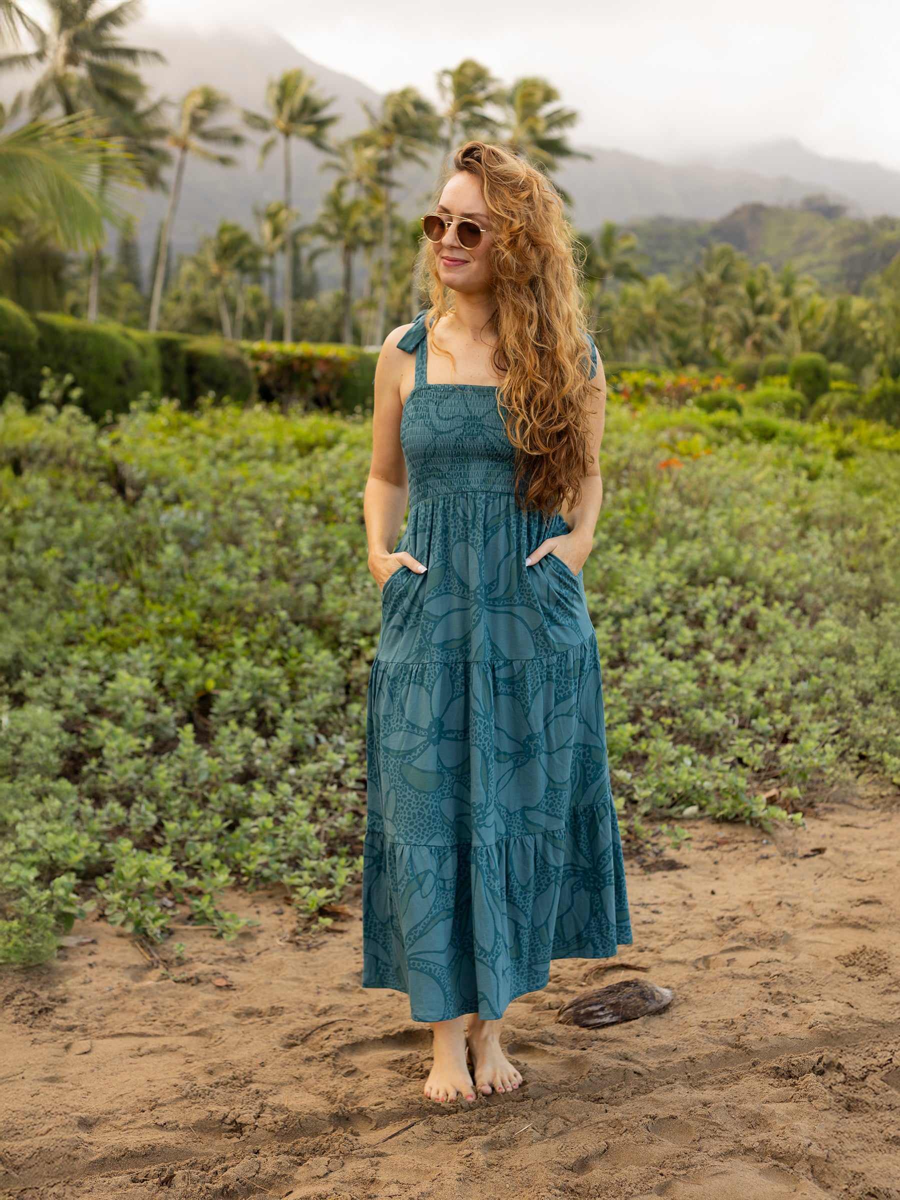 Woman in a teal Evie Smocked Dress with large floral print, smocked bodice, ribbon ties, and tiered maxi skirt.