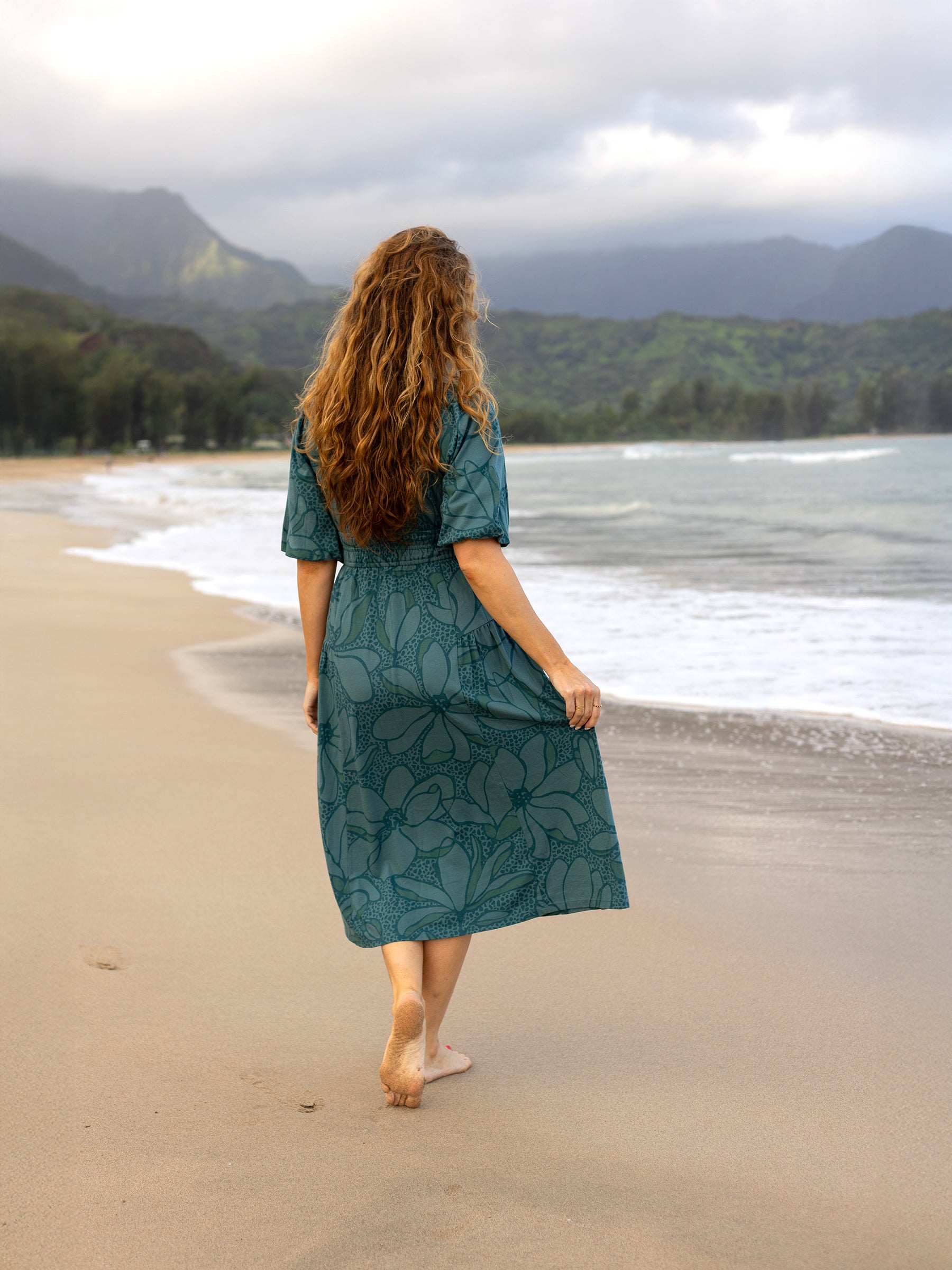 Woman in a teal Ophelia midi dress with large floral print, smocked waist, and puffed sleeves, walking barefoot on a beach.