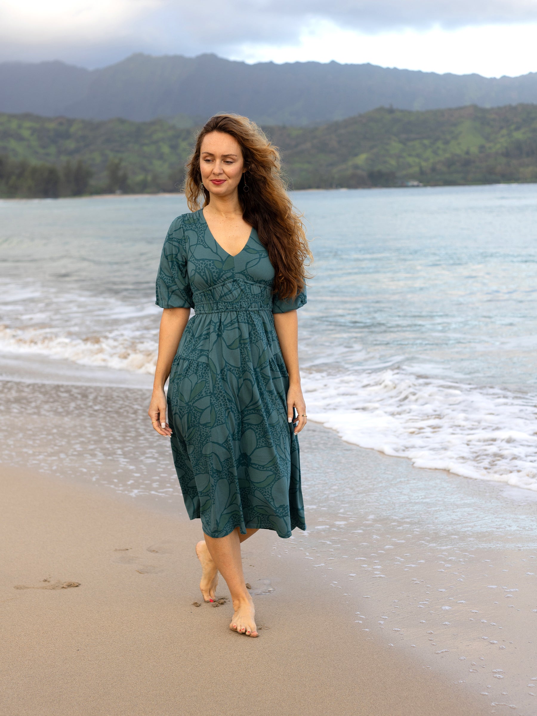Woman in a teal Ophelia midi dress with large florals, V-neck, puffed sleeves, and smocked waist, walking on a beach.