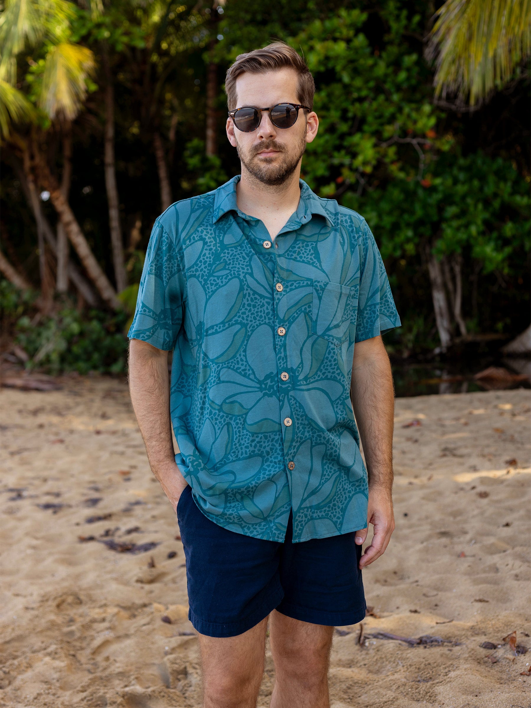 Man in Rincon Oasis teal organic cotton floral shirt with wooden buttons and navy shorts on a beach.