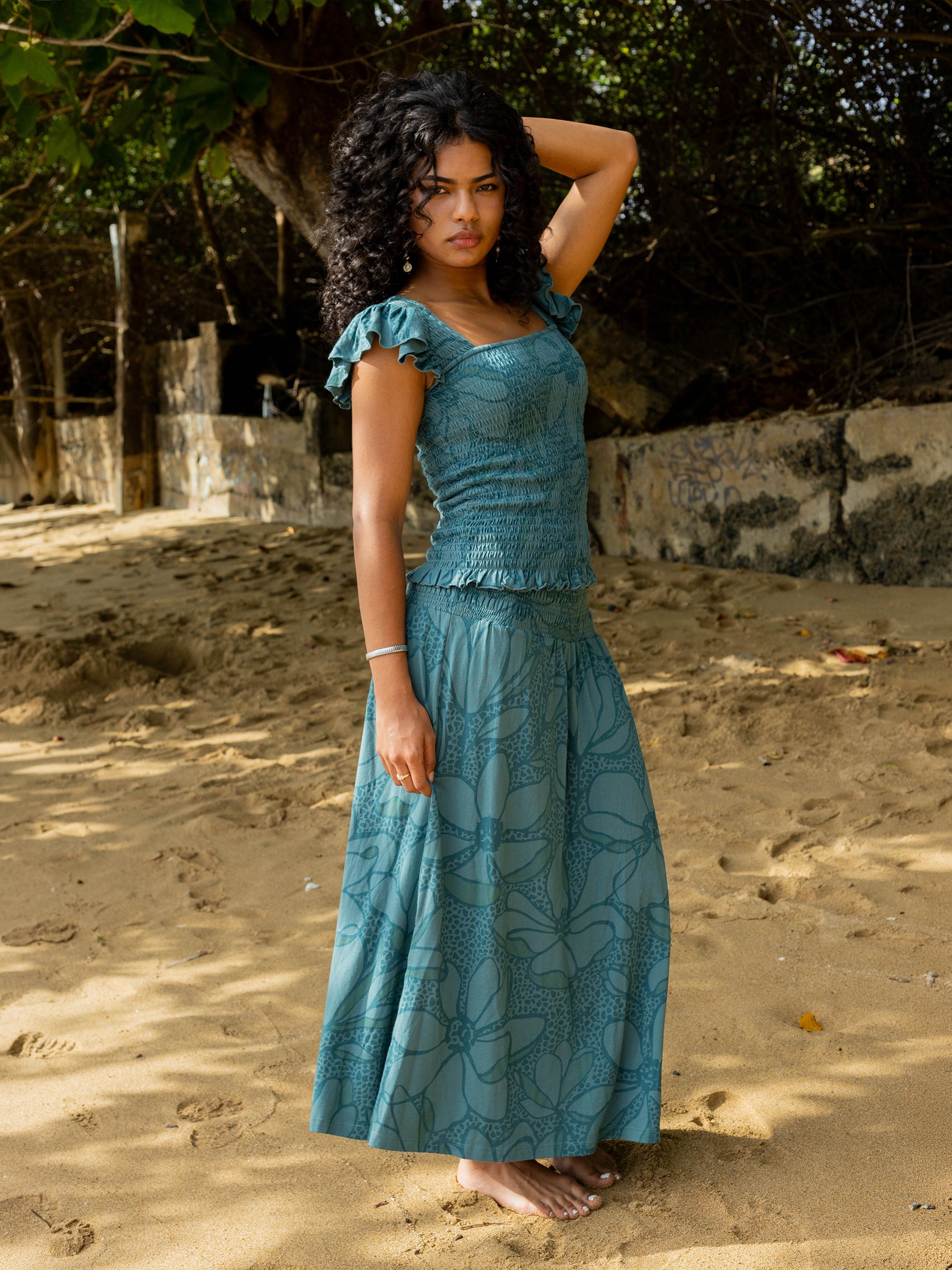 Woman in a seaside teal smocked maxi skirt with large florals and matching top on a sandy beach.