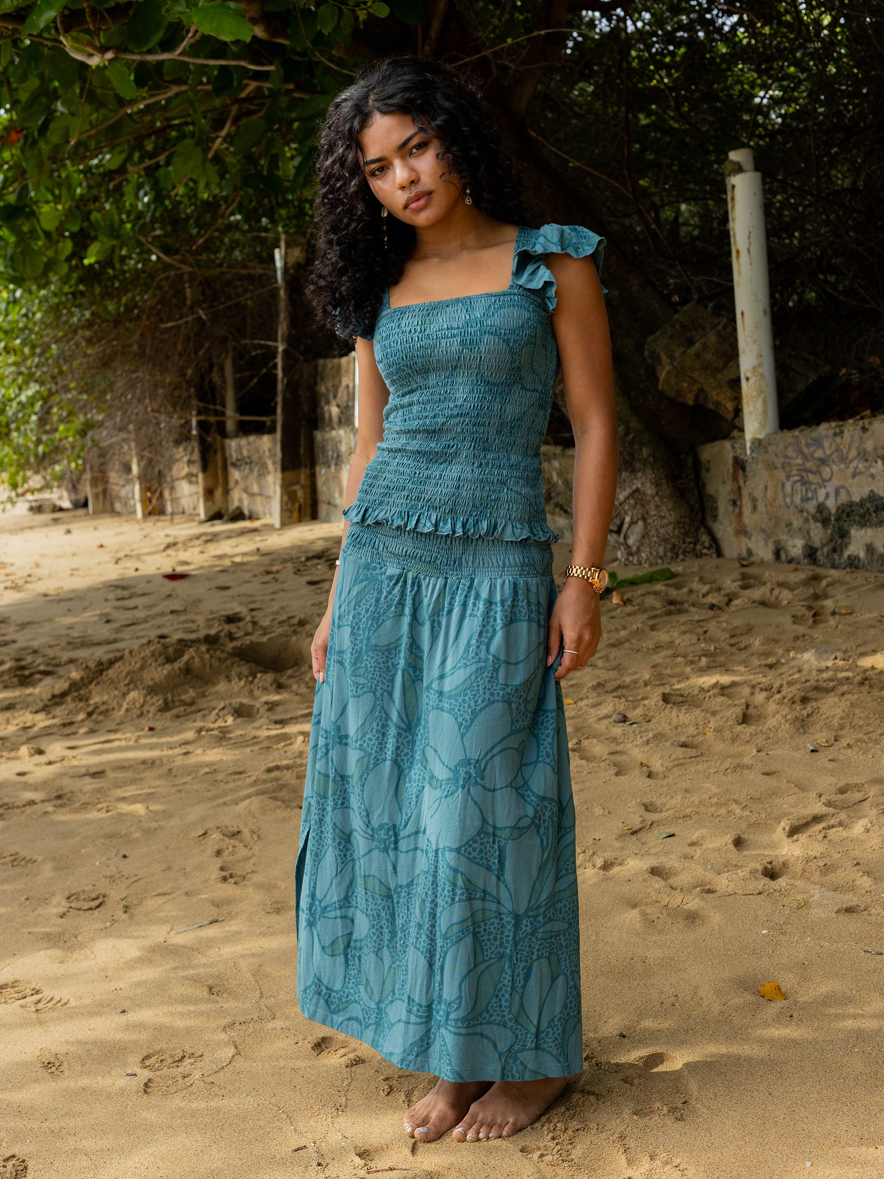 Woman in a teal smocked maxi skirt with floral print, side slit, and matching top on a sandy beach.