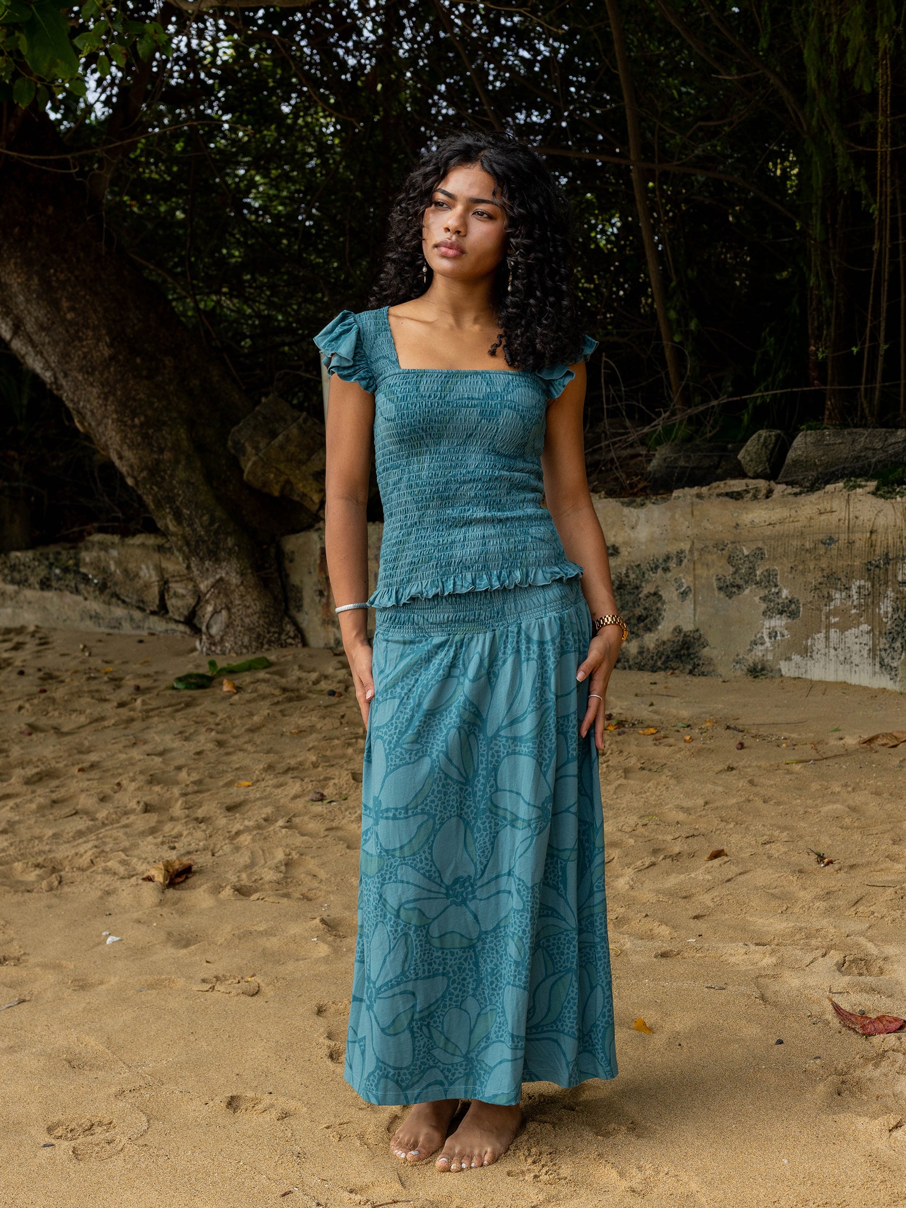 Woman in a teal smocked top with square neckline, flutter sleeves, and ruffled hem, paired with a matching floral maxi skirt on a beach.