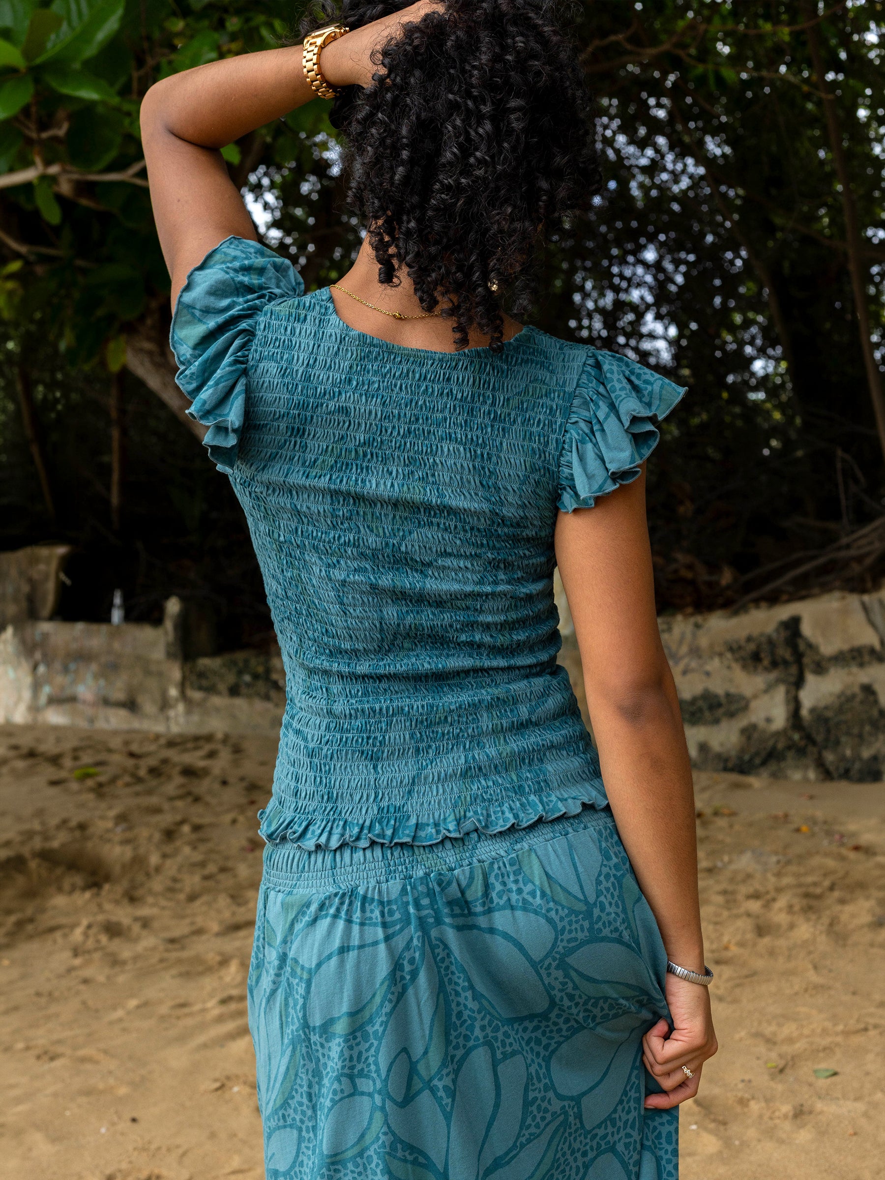 Woman from behind wearing a teal smocked top with flutter sleeves and a matching floral skirt on a beach.