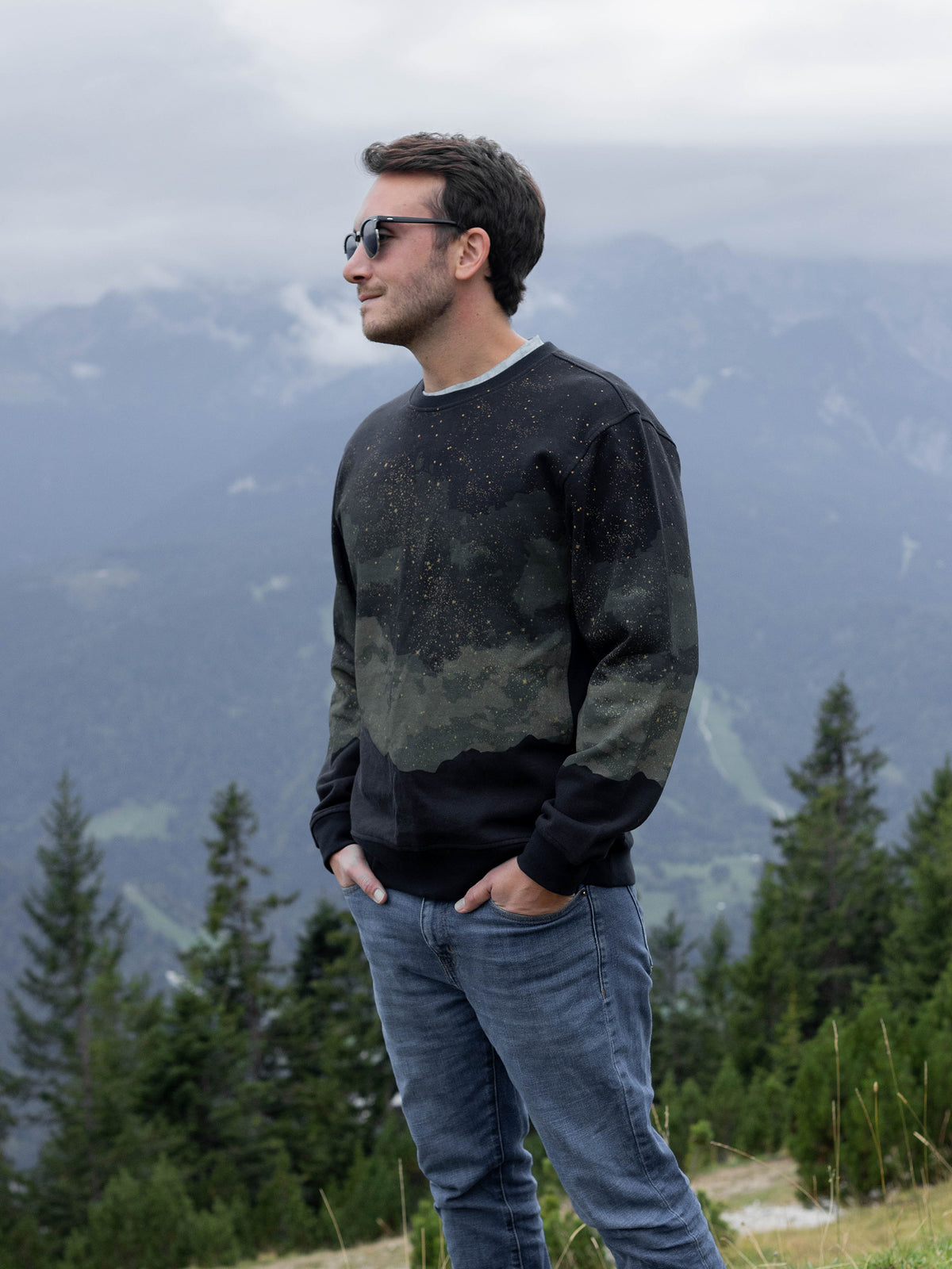 Model in sunglasses wearing the black Embercrest organic cotton sweatshirt with jeans on a mountainside